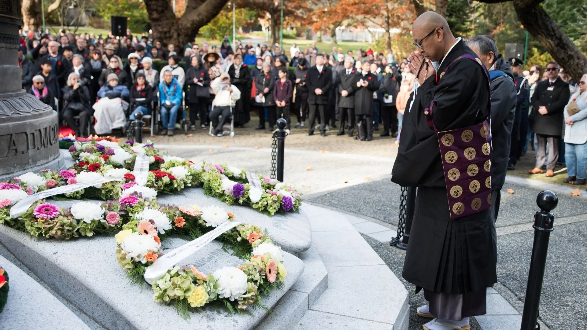 A person in traditional attire praying at a memorial with floral wreaths, a crowd watches in the background.