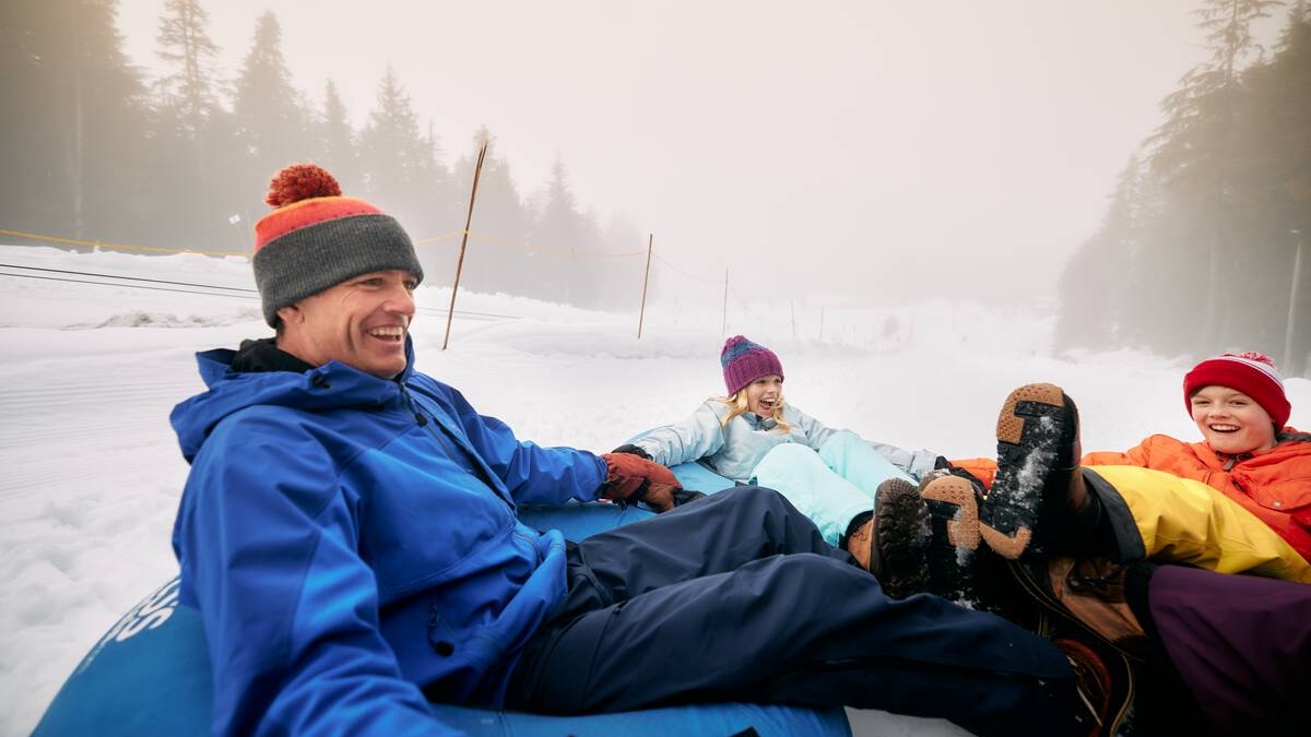 A father and two kids sit in snow tubes and hold hands as they zoom down a slope at the tube park at Cypress Mountain.