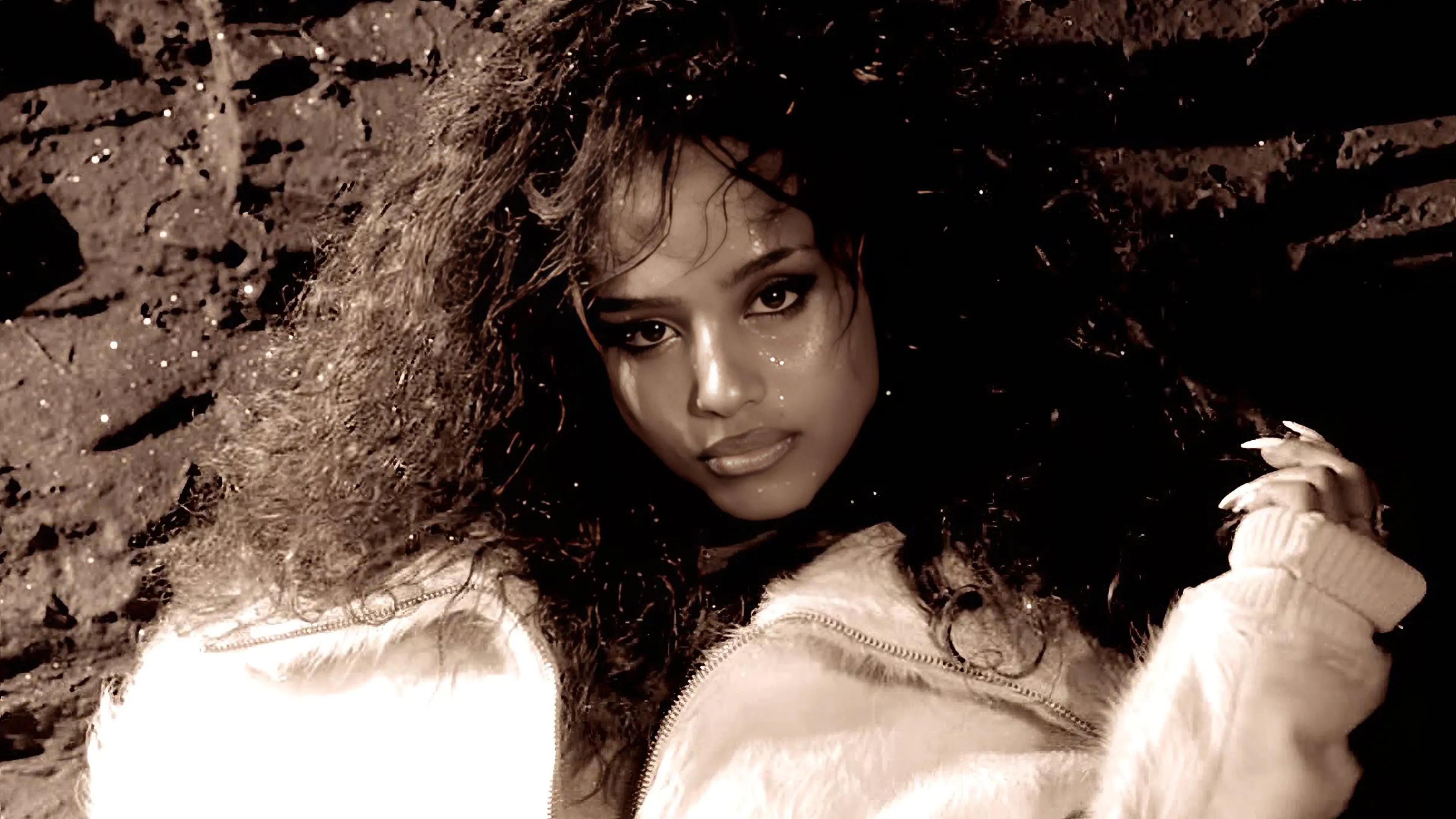 Person with long curly hair lying down, wearing a light-colored fuzzy jacket, looking directly at the camera.