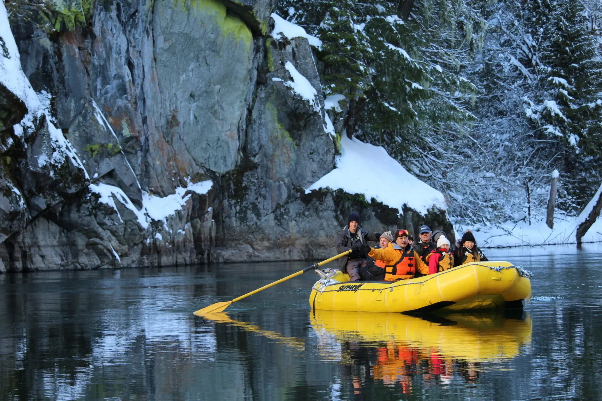 A group of people sit on a white water raft in a calm river with snow all around.
