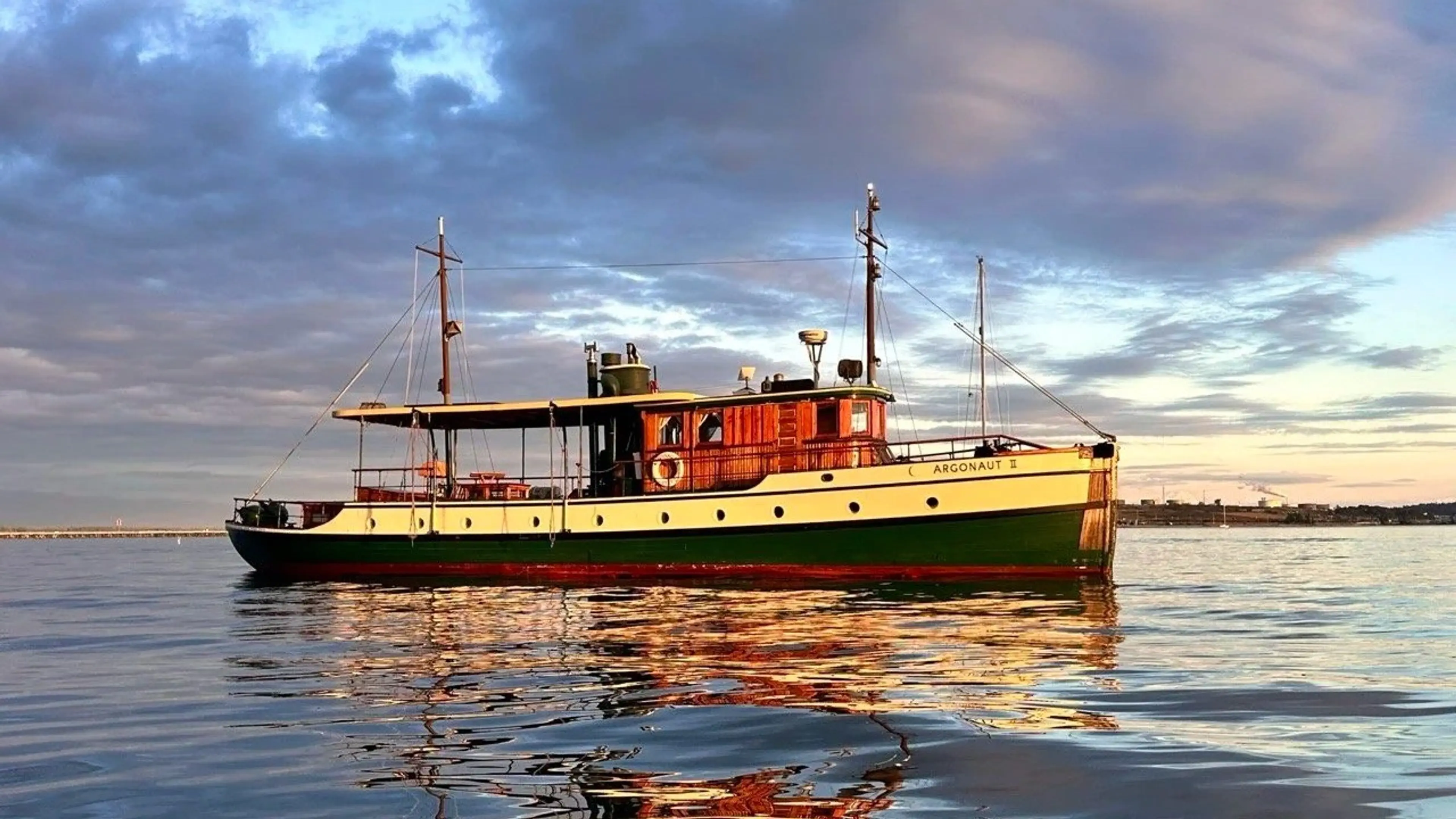 A vintage boat named Argonaut II floating on calm water under a partly cloudy sky at sunset.