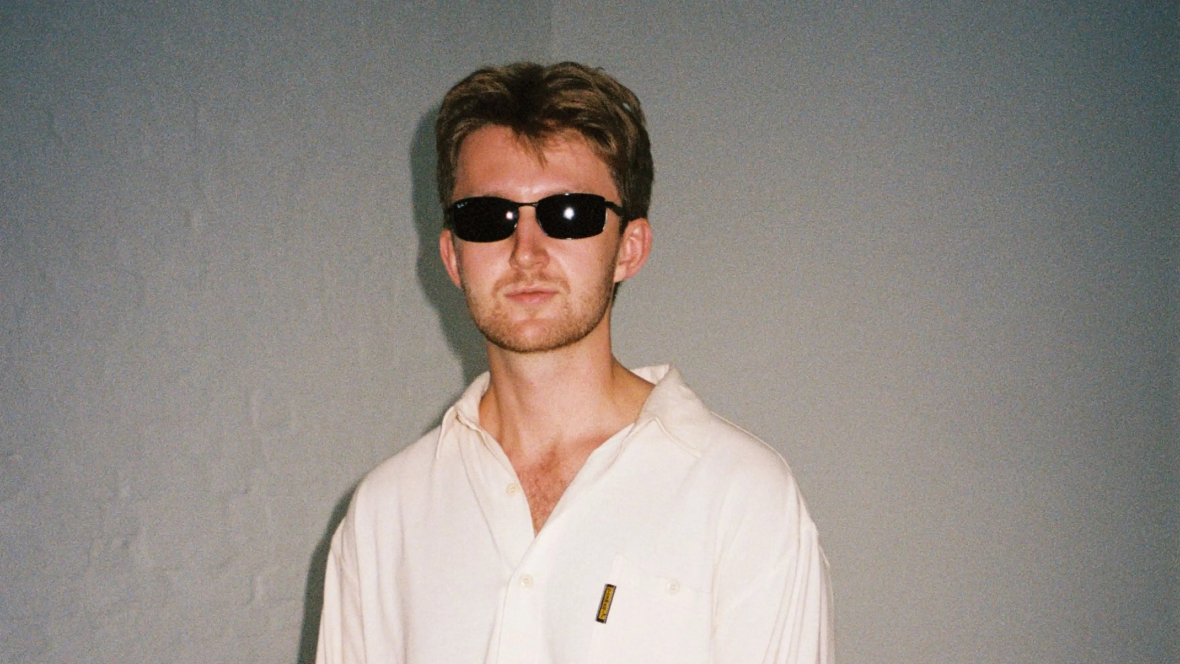 Person wearing black sunglasses and a white button-up shirt standing against a plain gray wall.