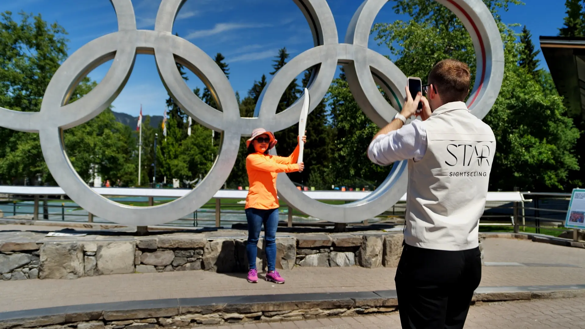Person in a jacket posing with a torch in front of large Olympic rings while another person takes a photo.