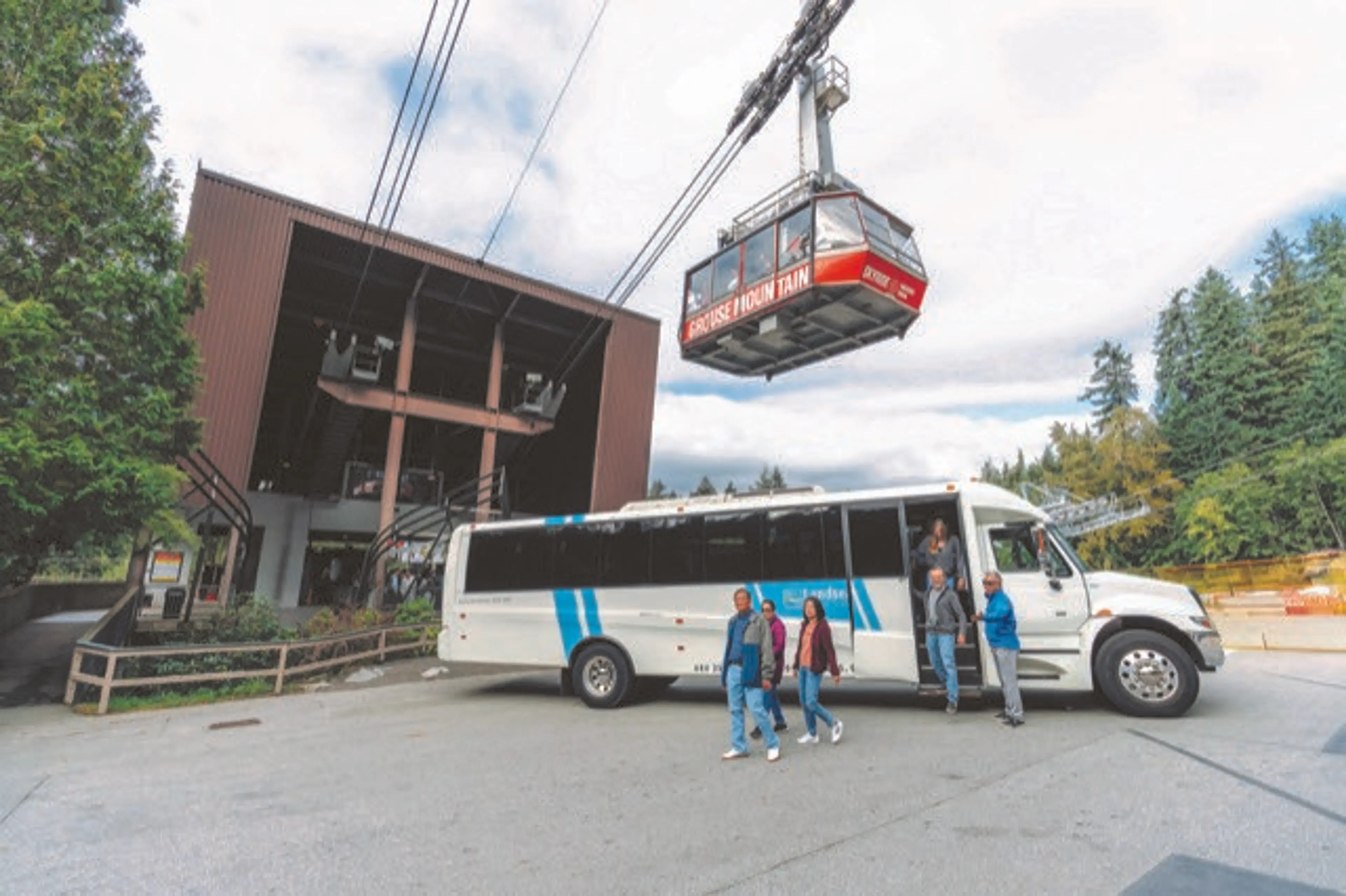 Five people walking near a white shuttle bus parked outside a cable car station with a red cable car labeled 'Grouse Mountain' suspended above.