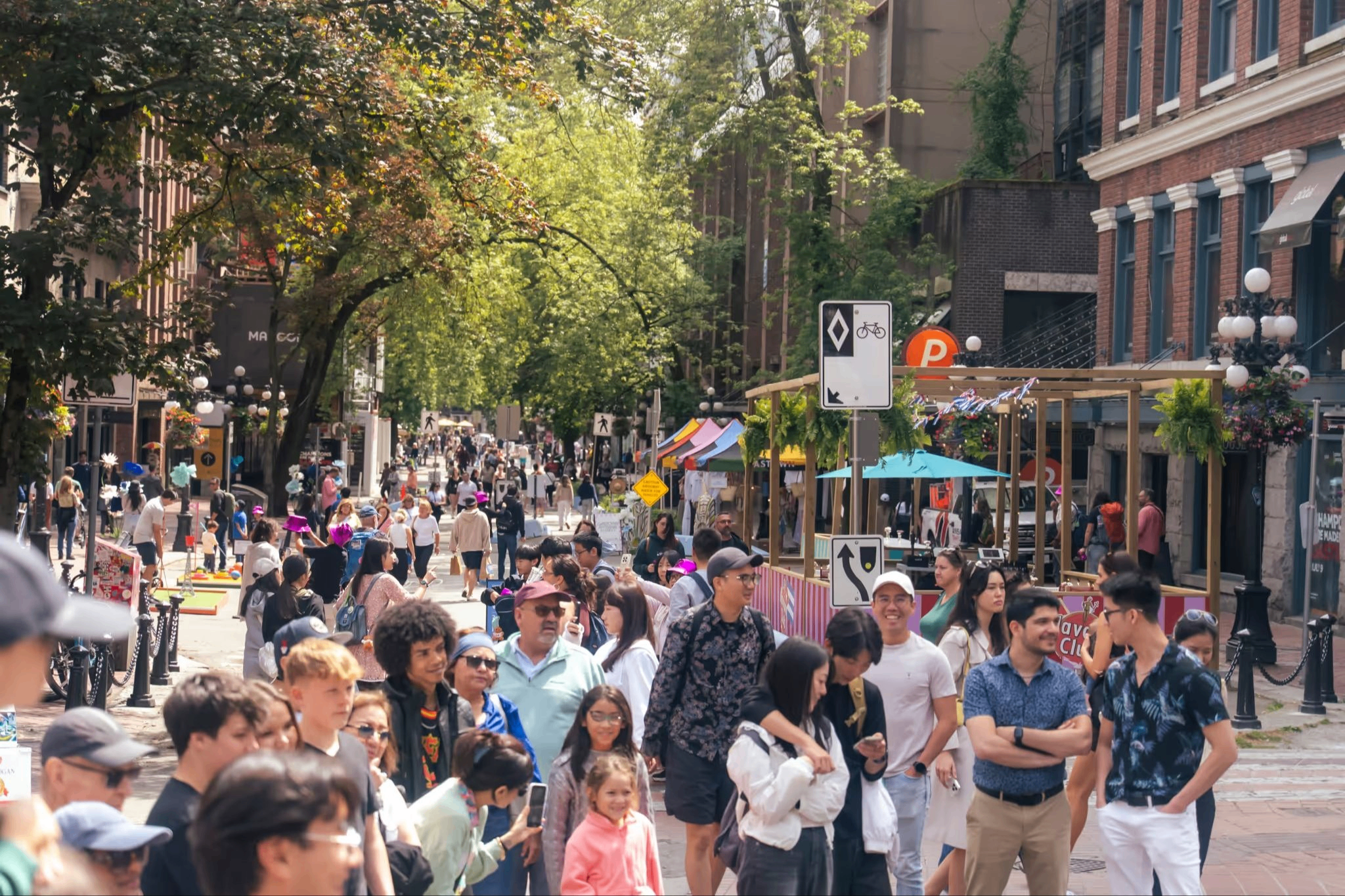 Crowded urban street with diverse people walking and socializing under green trees and near brick buildings.