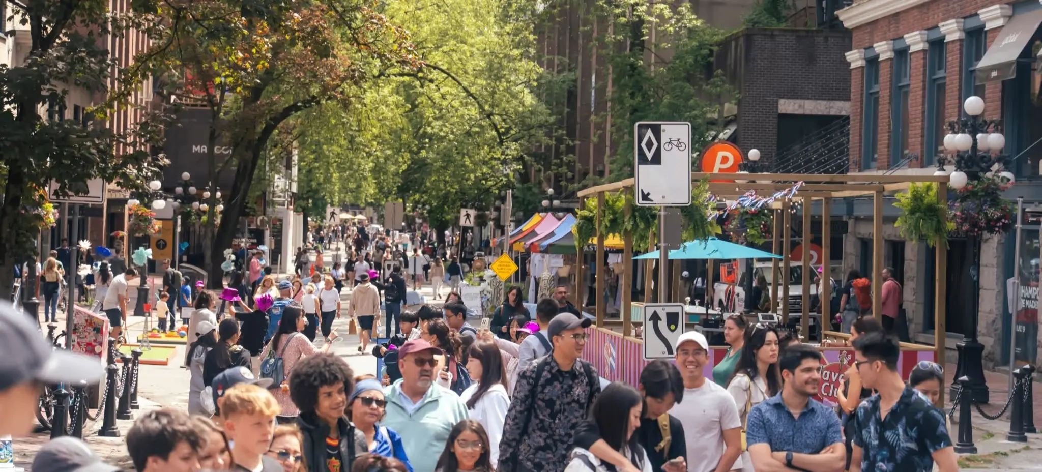 Crowded urban street with diverse people walking and socializing under green trees and near brick buildings.
