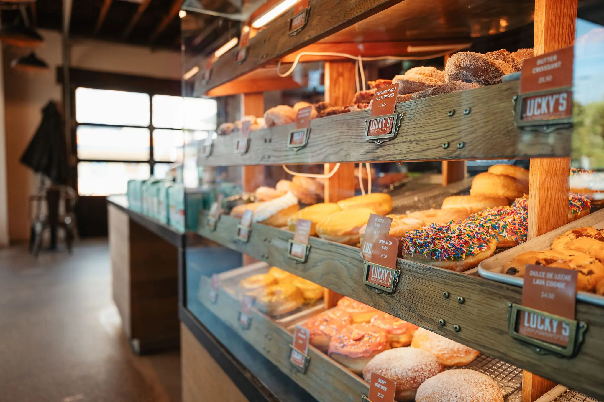 Wooden shelves displaying various donuts and pastries with price tags inside a bakery.