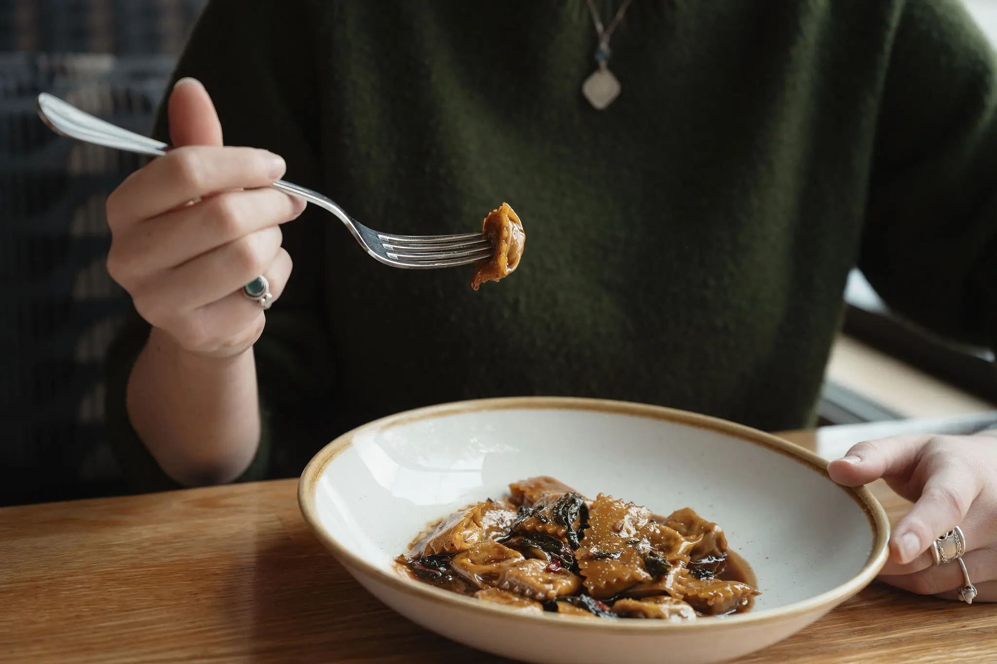 Person holding a fork with a piece of ravioli above a bowl of ravioli in sauce on a wooden table.