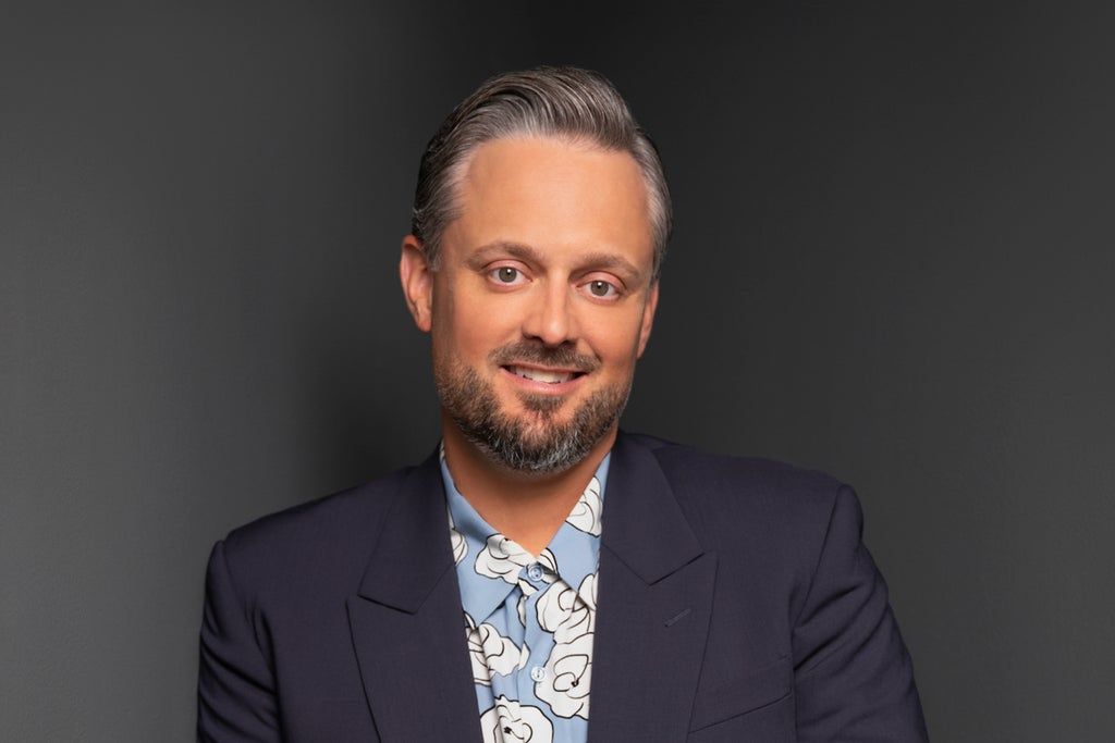 Person with short slicked-back hair and beard wearing a dark blazer over a blue shirt with white floral patterns, smiling against a dark background.