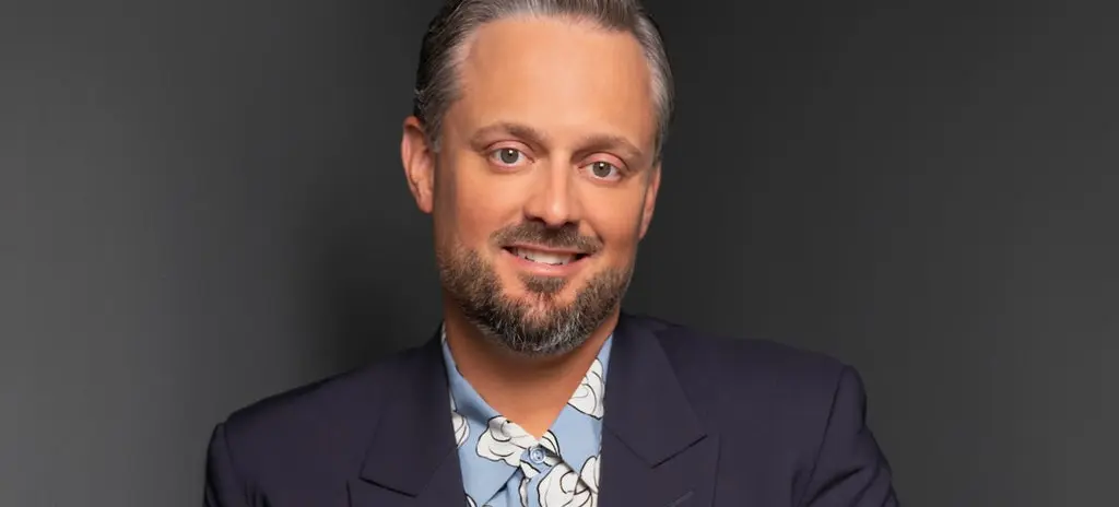Person with short slicked-back hair and beard wearing a dark blazer over a blue shirt with white floral patterns, smiling against a dark background.