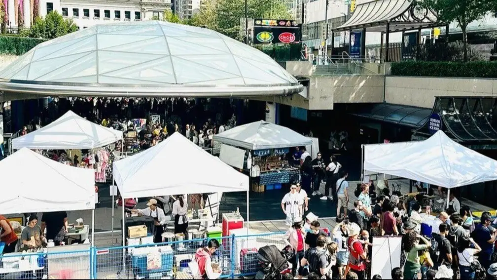 Outdoor market with white tents and many people browsing and walking around in a city setting.