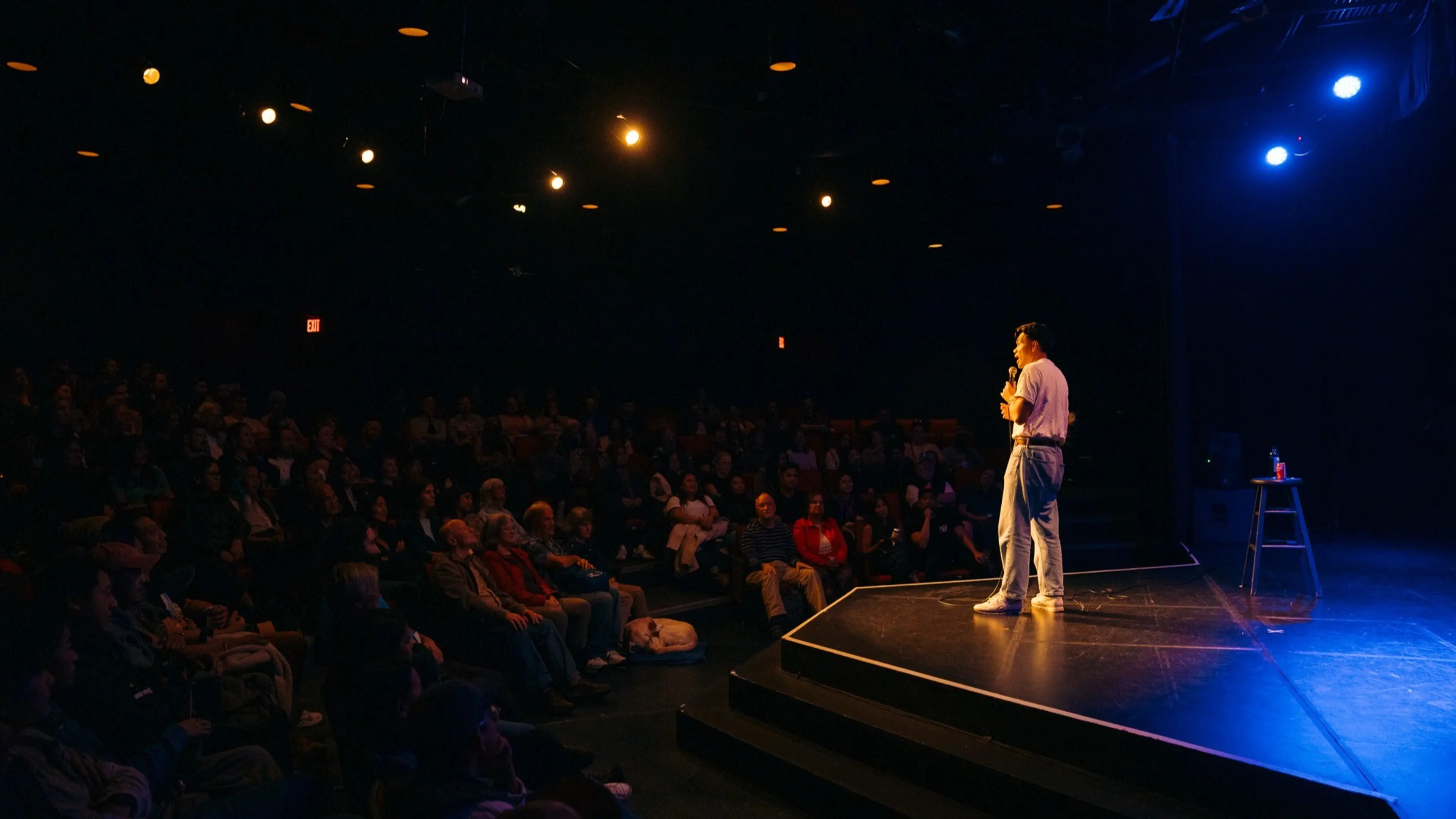 A person stands on a stage with a microphone speaking to a seated audience in a dimly lit theater.