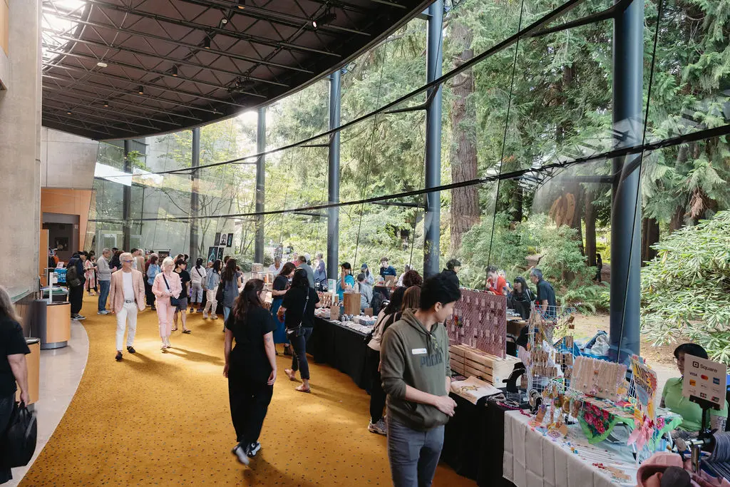 People browsing and shopping at the halls at the Come Towards the Fire event with large glass windows showing trees outside.