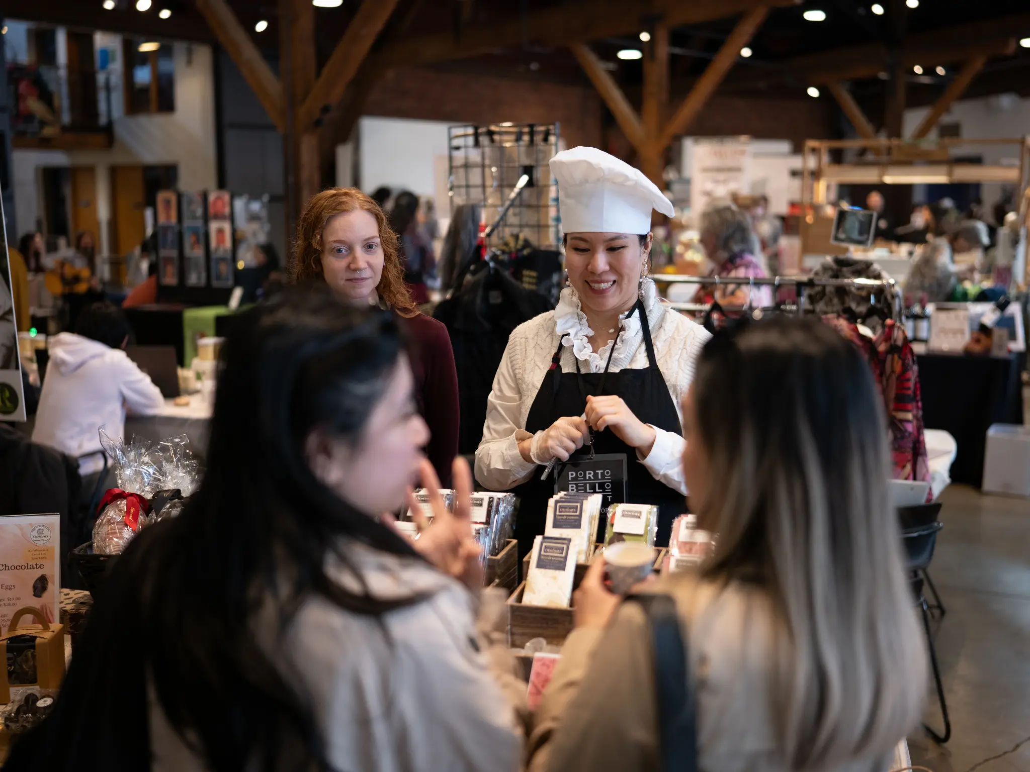 A person in a chef hat and apron smiles behind a market stall with packaged goods, while three people stand in front.