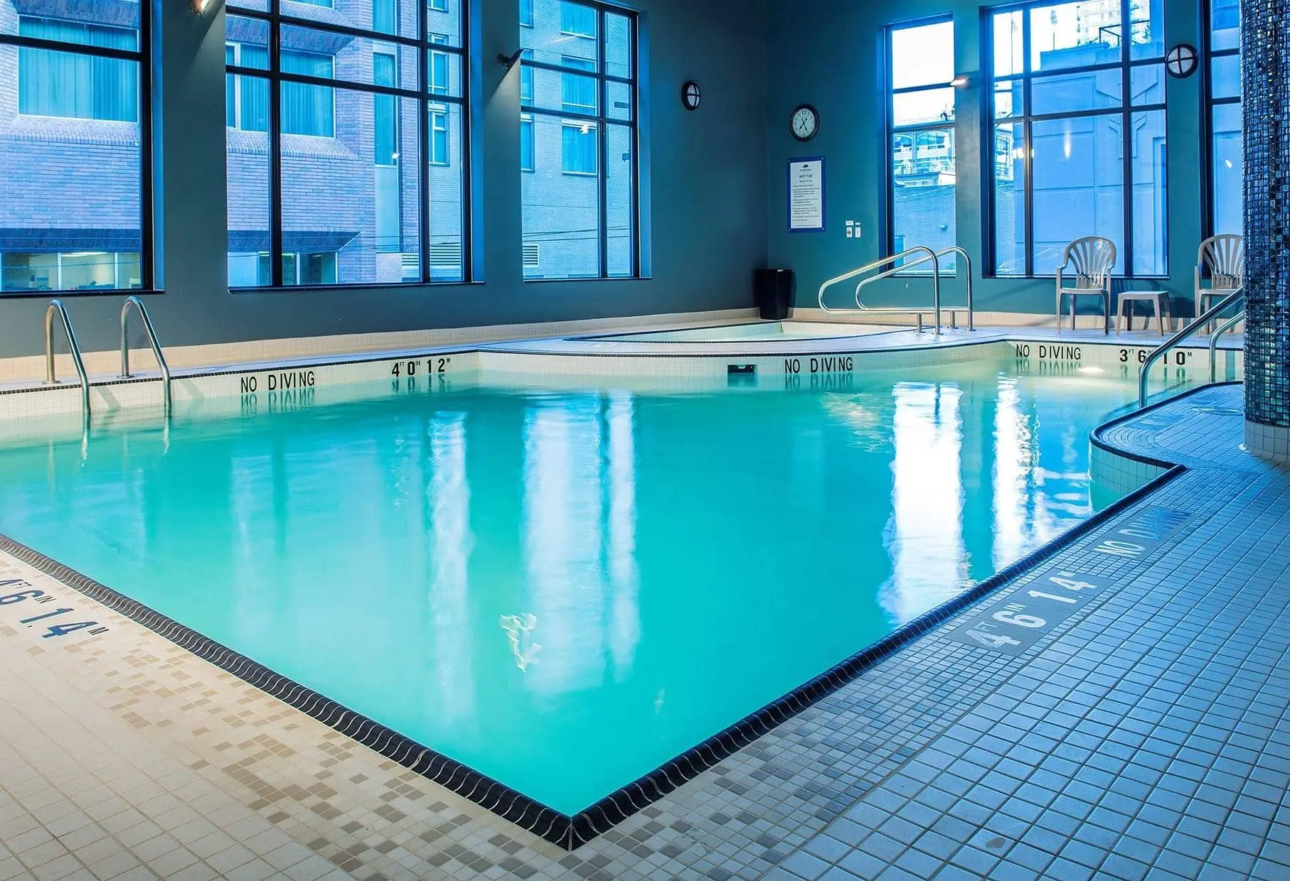 A calm, empty indoor pool with blue water, white tiles, and chairs sitting pool-side.
