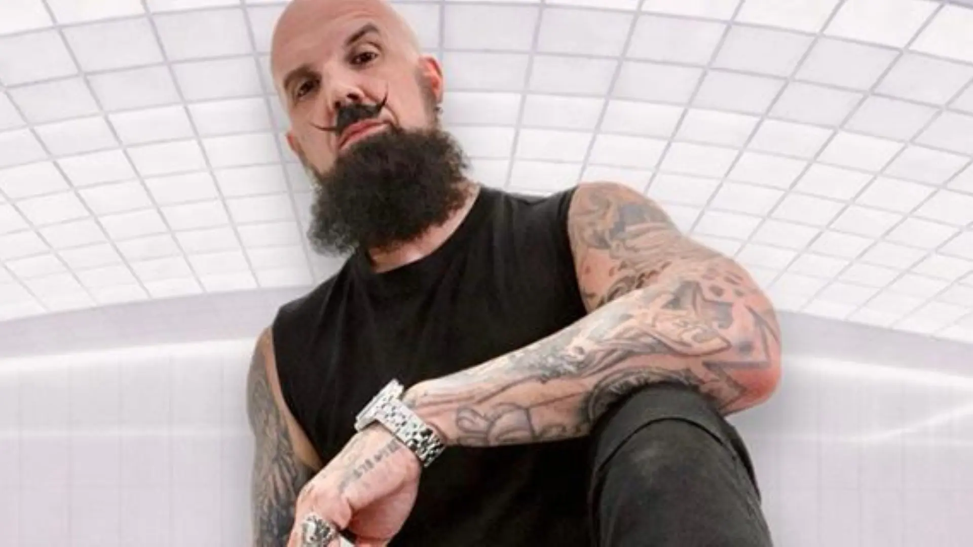 Person with a bald head, styled mustache, and full beard wearing a black sleeveless shirt, showing tattooed arms and a watch.