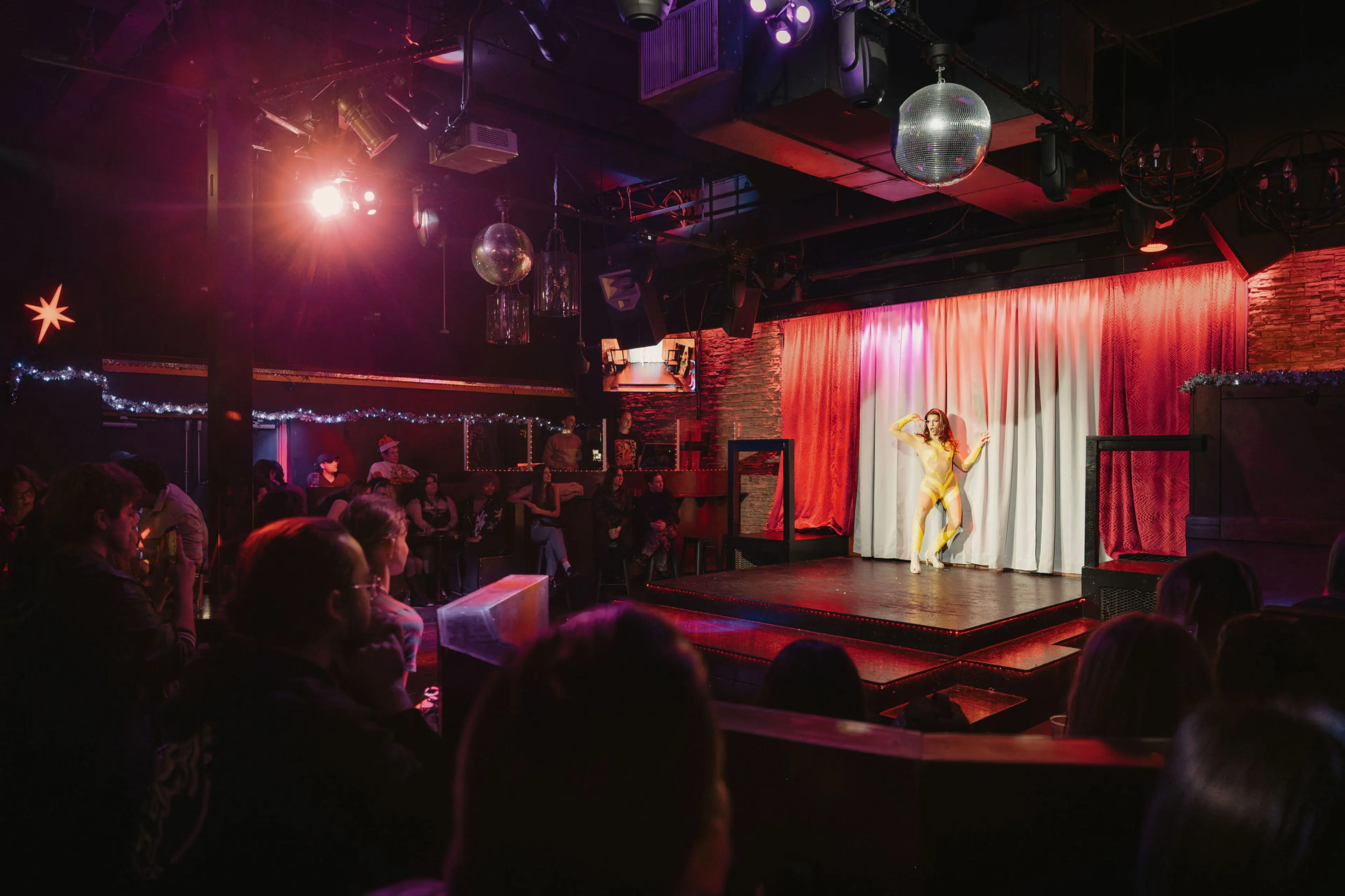 A person in a yellow outfit dancing on a small stage with red and white curtains, watched by an audience in a dimly lit club.