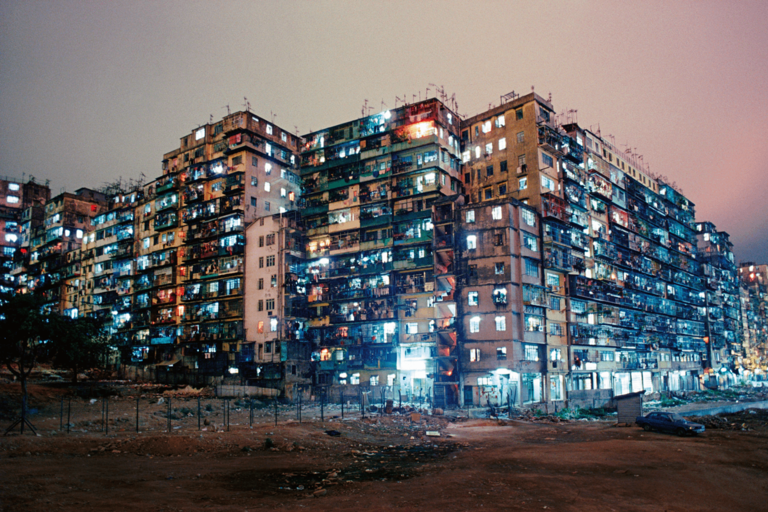 Large, densely packed apartment buildings at night with many illuminated windows and a dirt foreground.