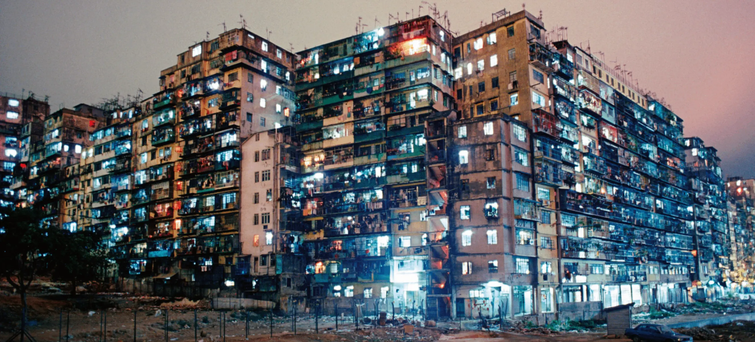 Large, densely packed apartment buildings at night with many illuminated windows and a dirt foreground.