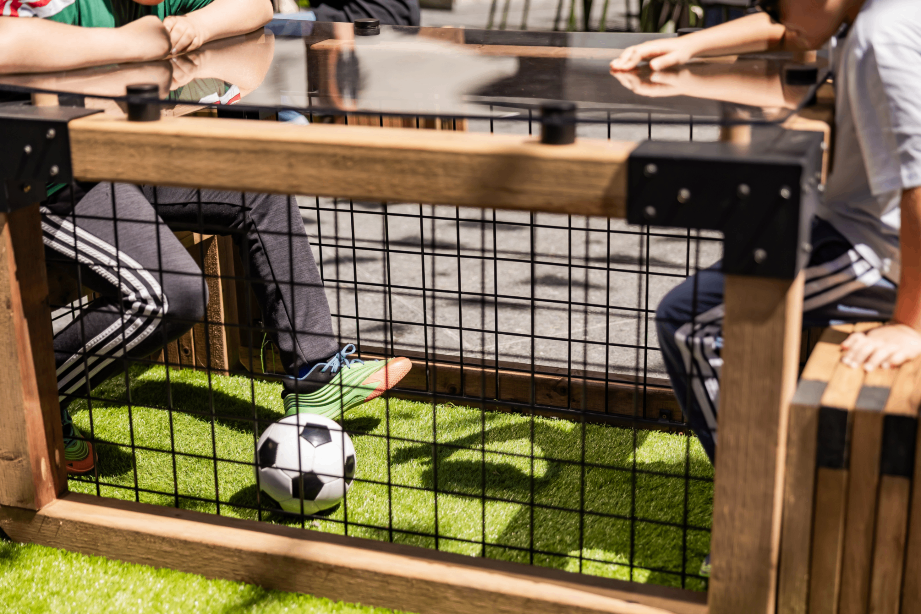 Two people sitting at a wooden table with a metal grid, playing foot soccer with a small black and white ball on artificial grass.