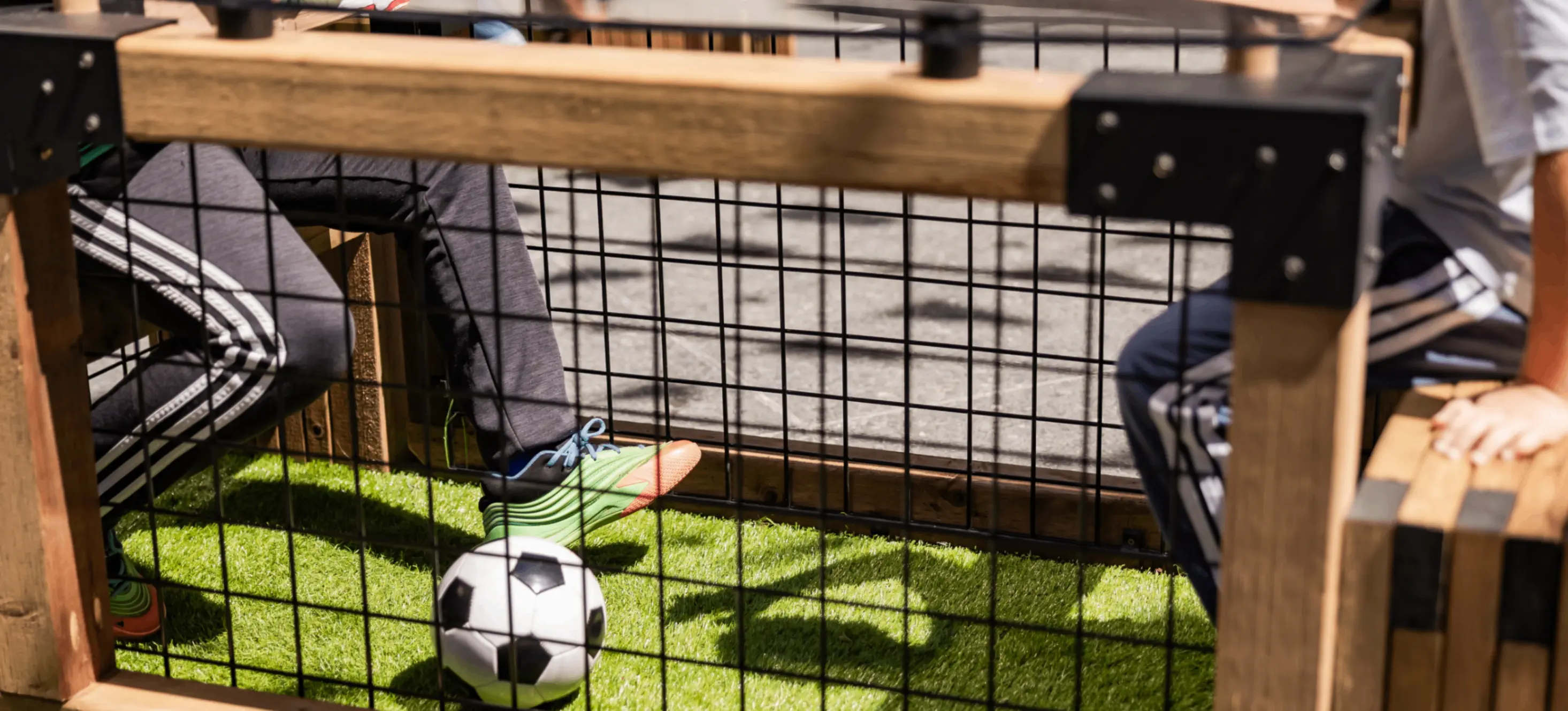 Two people sitting at a wooden table with a metal grid, playing foot soccer with a small black and white ball on artificial grass.