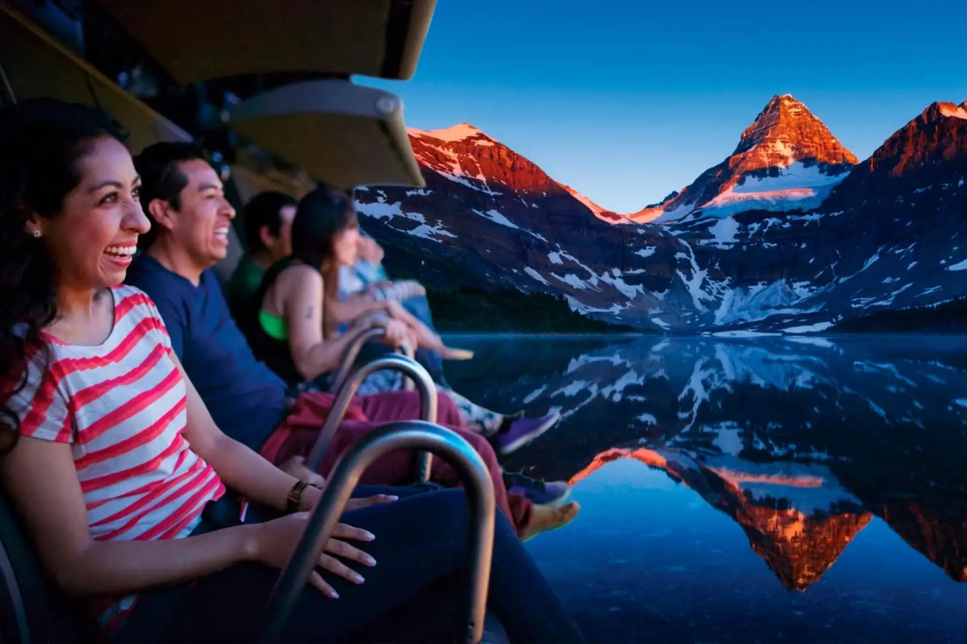 Group of people sitting and smiling on a ride at Flyover Canada looking at visualization of snow-capped mountains and their reflection in calm water at sunset.