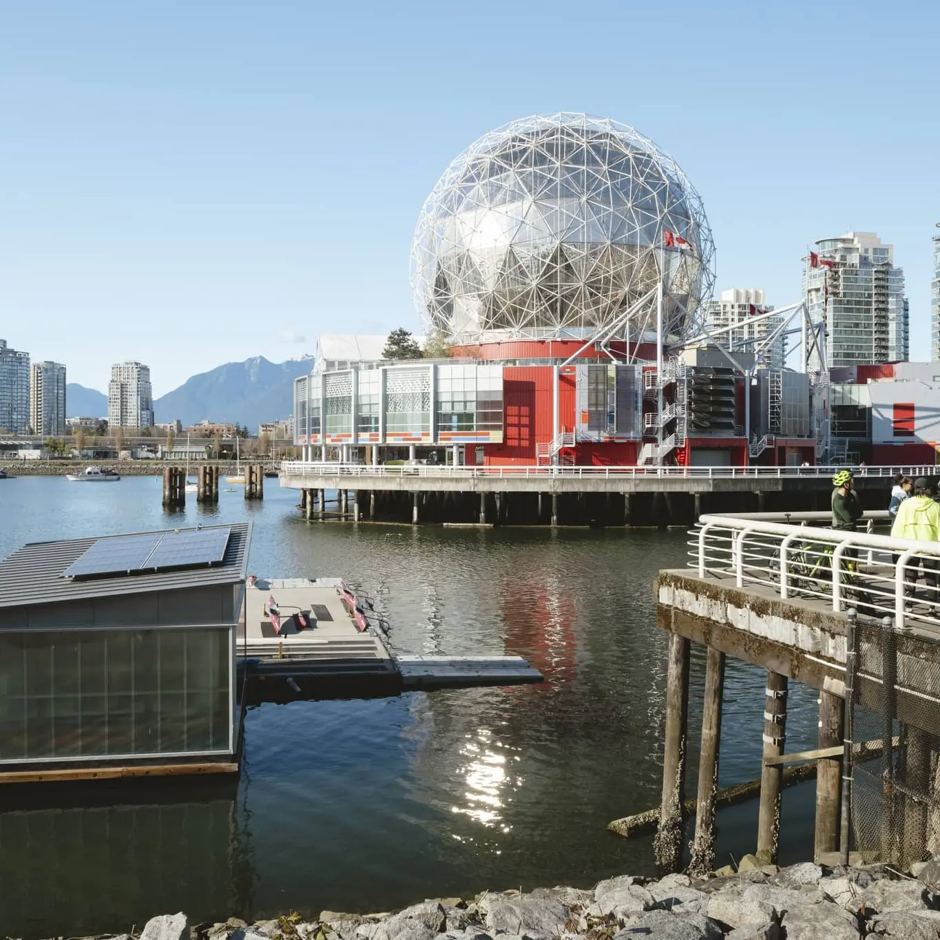 Waterfront scene with a geodesic dome building, modern high-rises, and people standing on a pier.