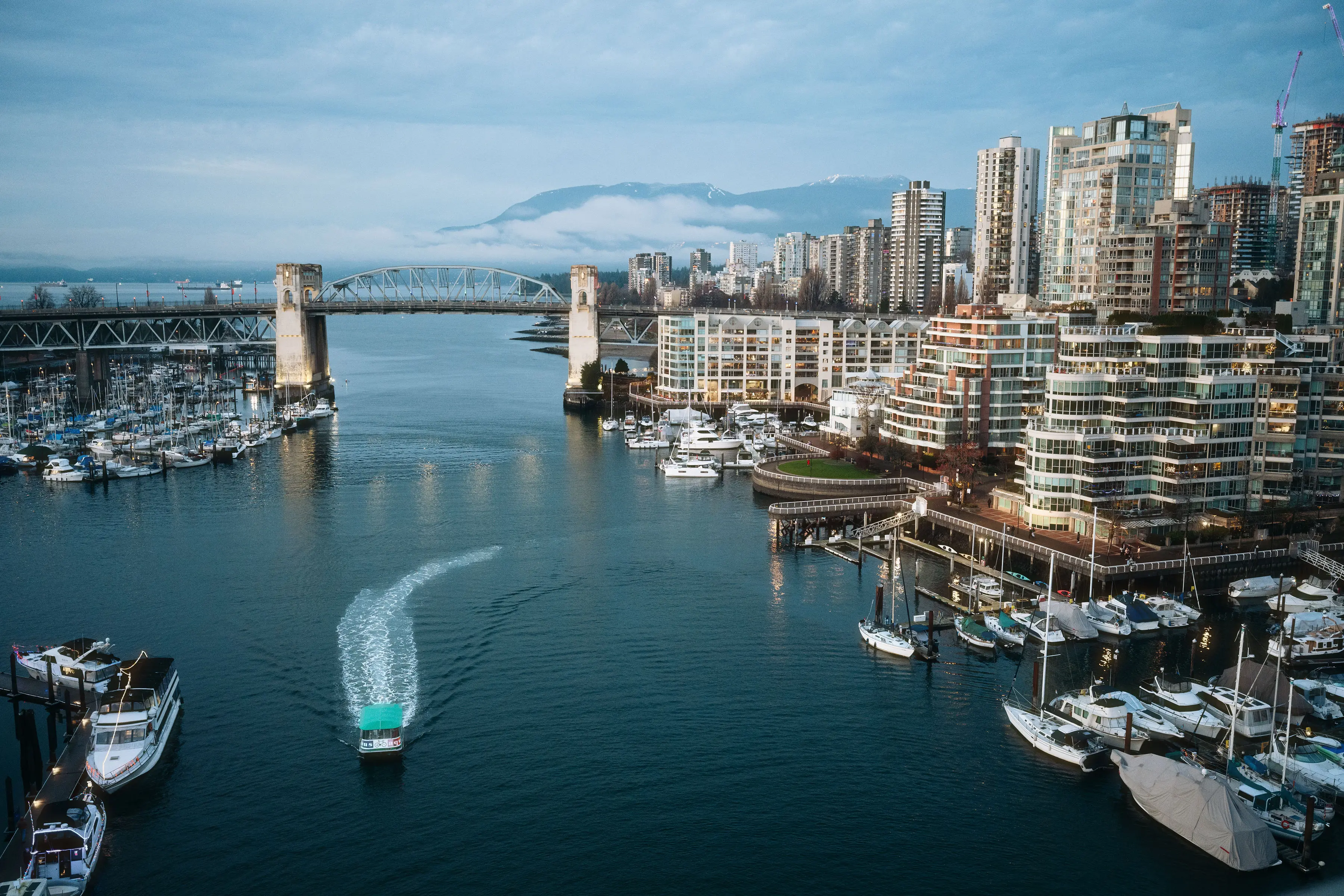 Boat moving through a waterway with a bridge, marina with boats, and city buildings under a cloudy sky.