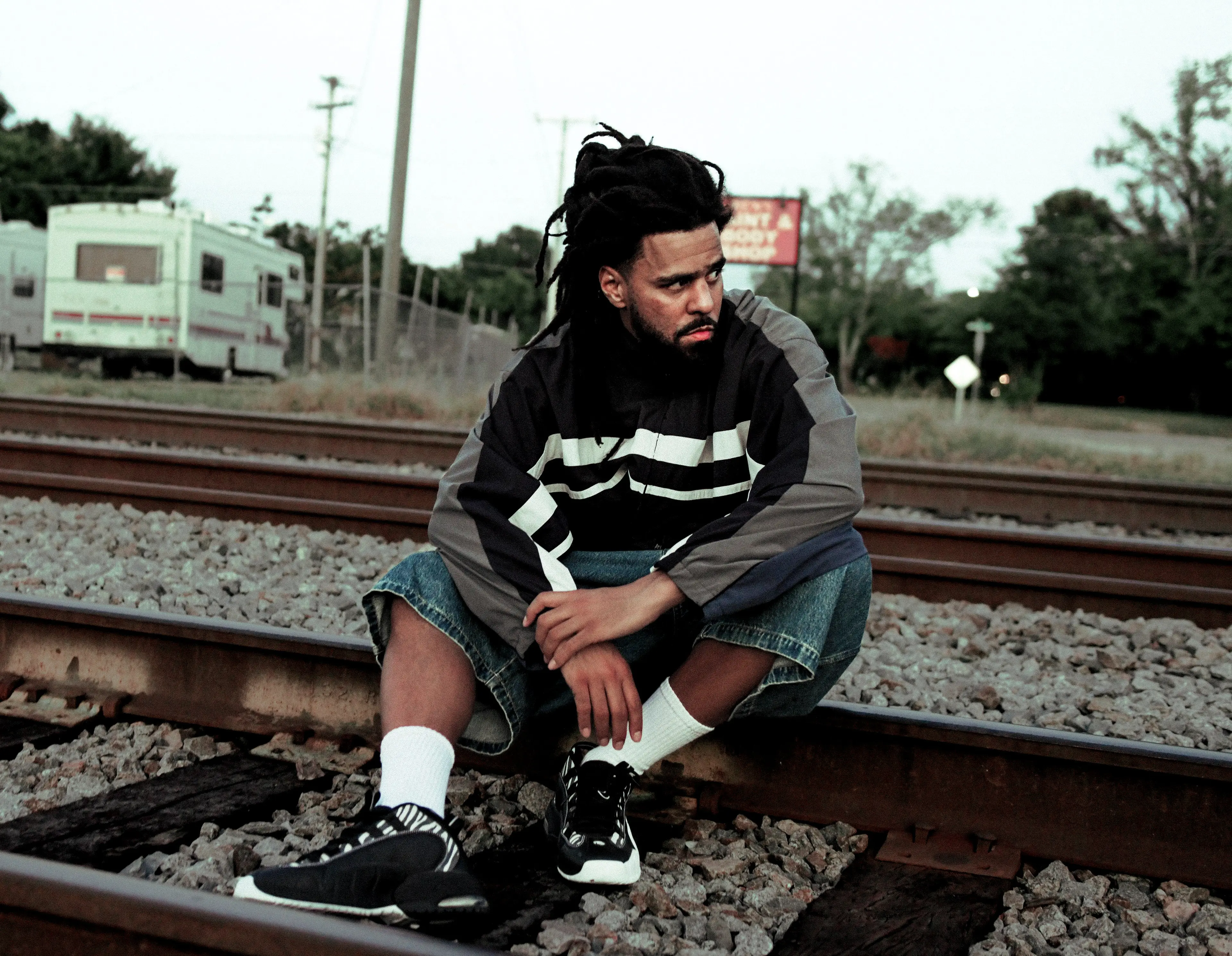 Person with dreadlocks sitting on railroad tracks wearing a black and gray jacket, denim shorts, white socks, and black sneakers.