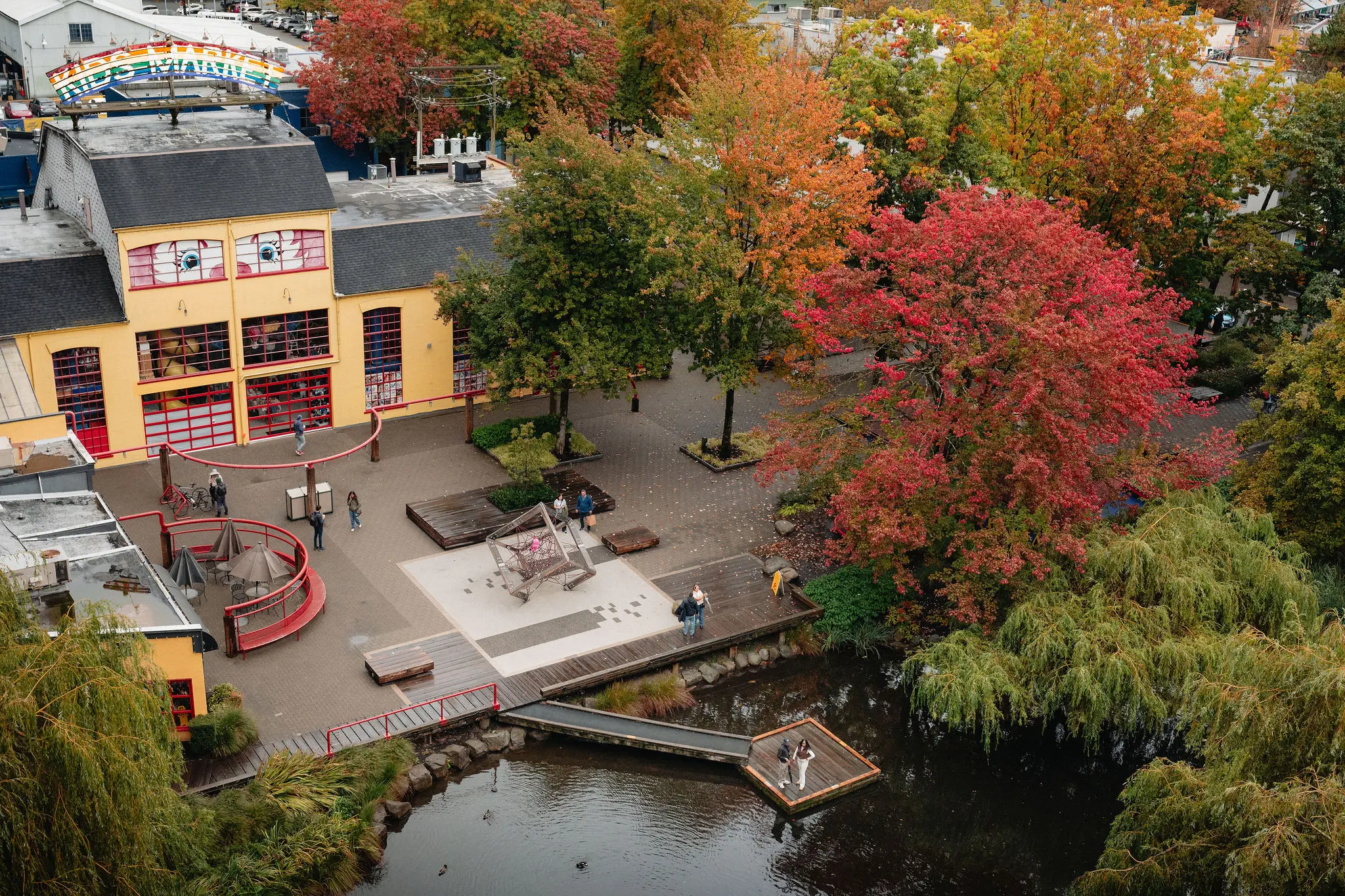 A colorful building with a rainbow sign reading 'Kids Market' near a pond surrounded by trees with autumn foliage and a small dock with two people.