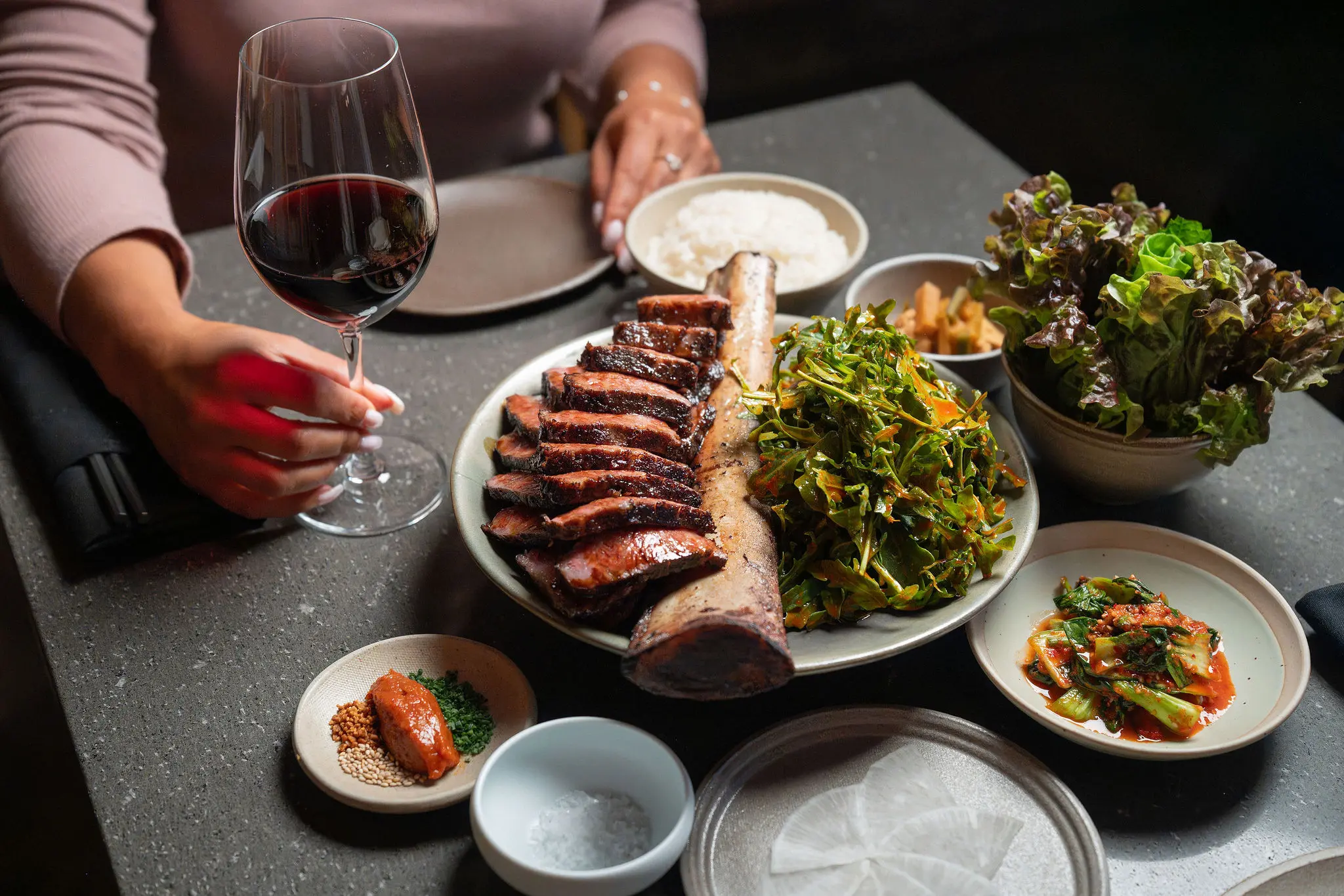 Person holding a glass of red wine next to a table with grilled meat on a bone, leafy greens, rice, kimchi, and various side dishes.