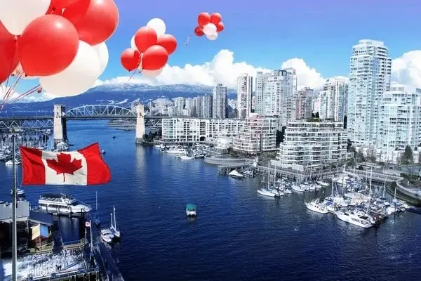 Red and white balloons and a Canadian flag over a harbor with boats and a city skyline with a bridge and mountains in the background.