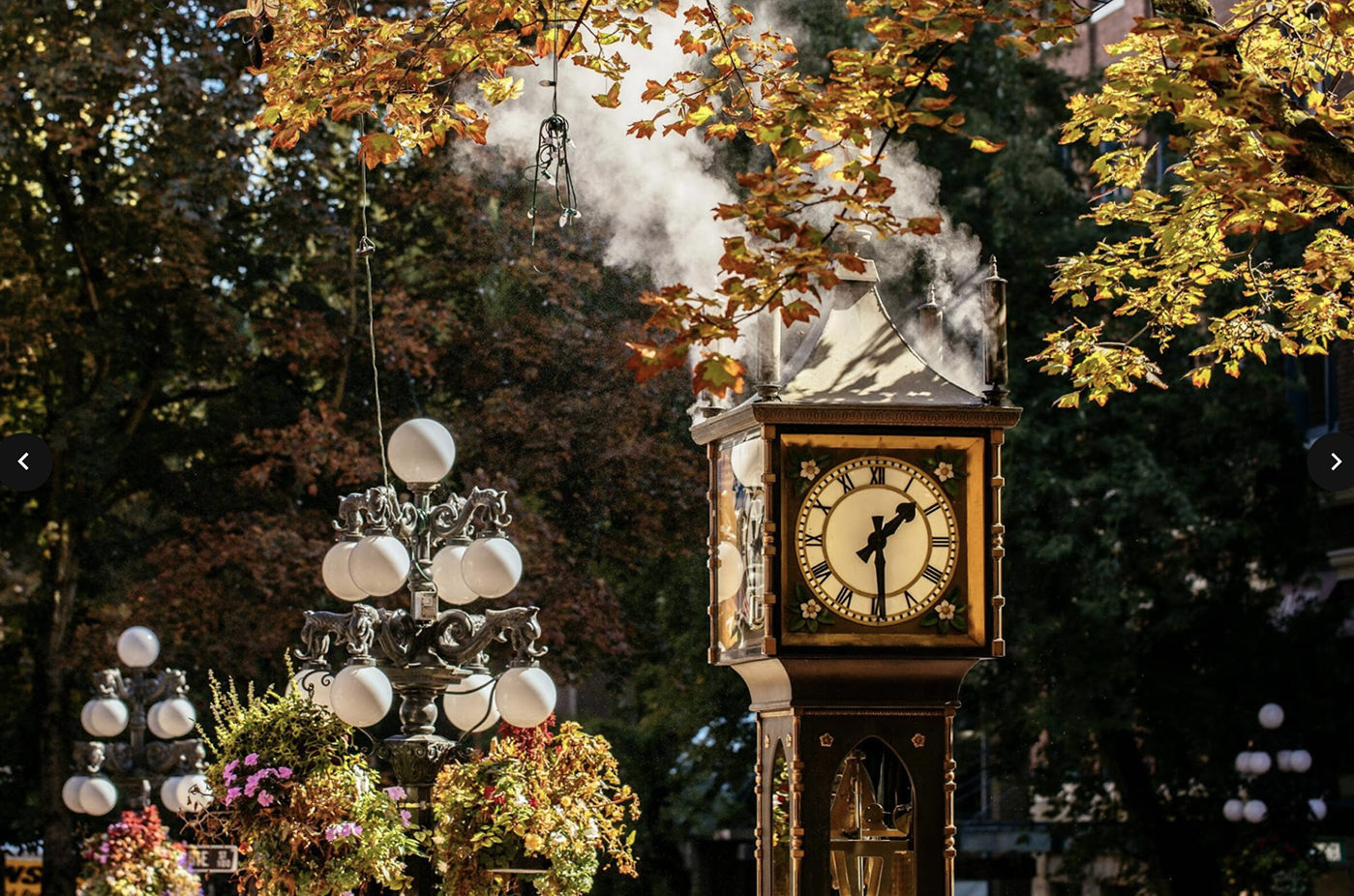 Steam clock showing 3:30 surrounded by autumn trees and vintage street lamps with hanging flower baskets.