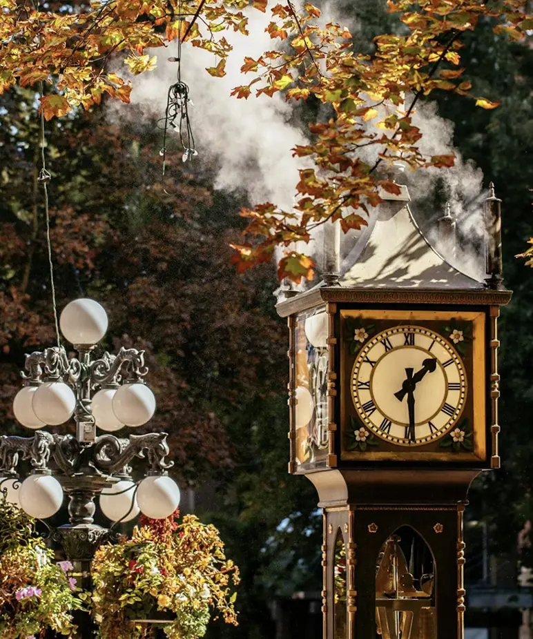 Steam clock showing 3:30 surrounded by autumn trees and vintage street lamps with hanging flower baskets.