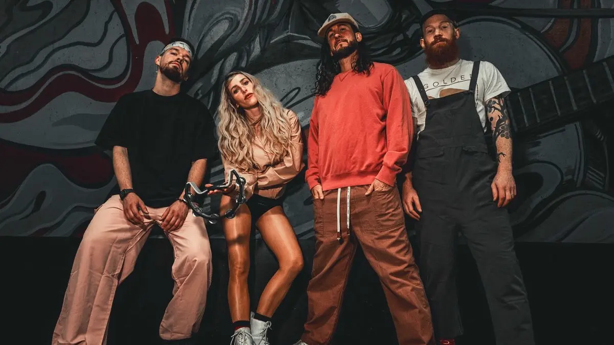 Four people standing in front of a graffiti wall, one holding a tambourine, dressed in casual streetwear.