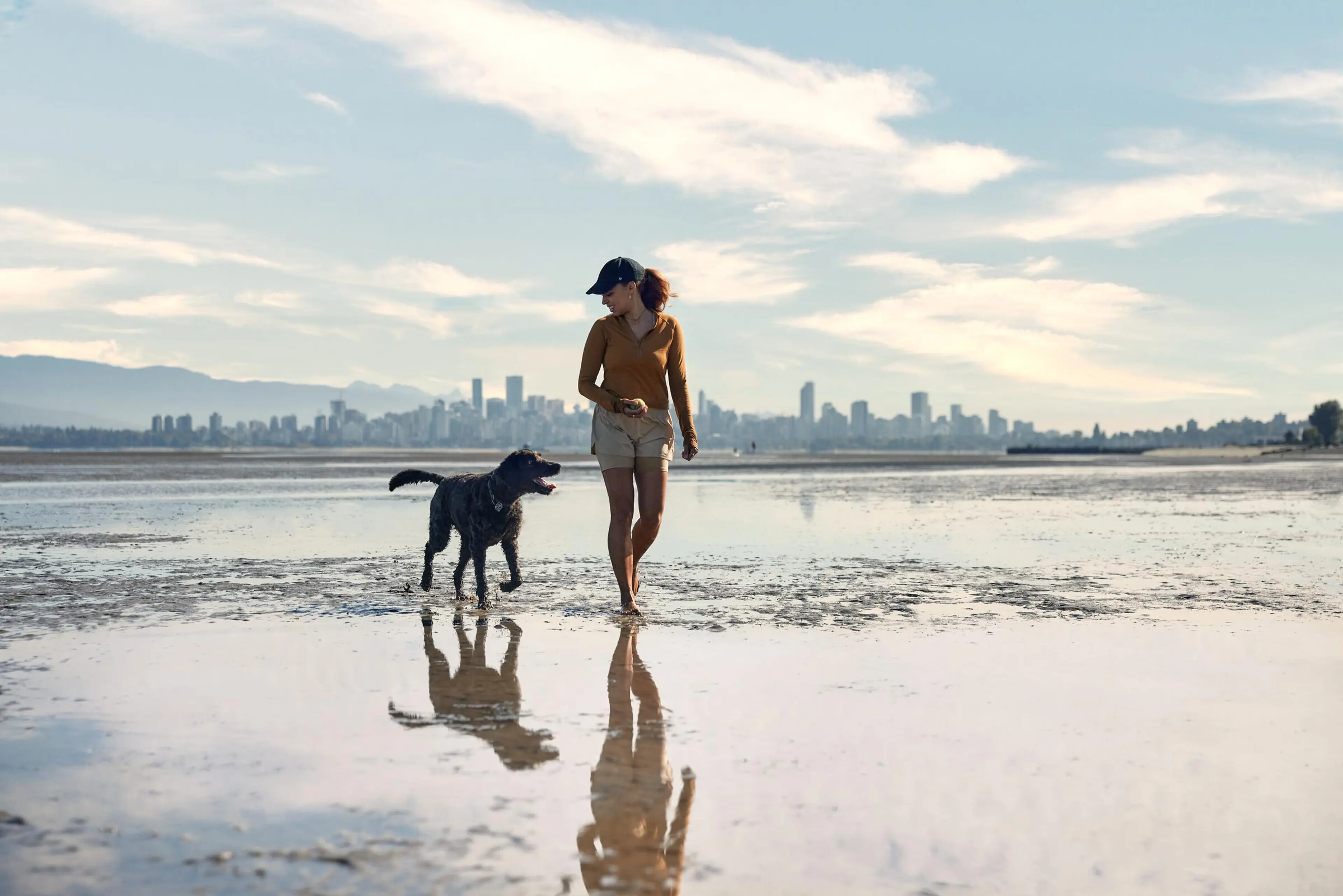Woman in shorts and cap walking barefoot with a black dog on a wet beach with city skyline in the background.
