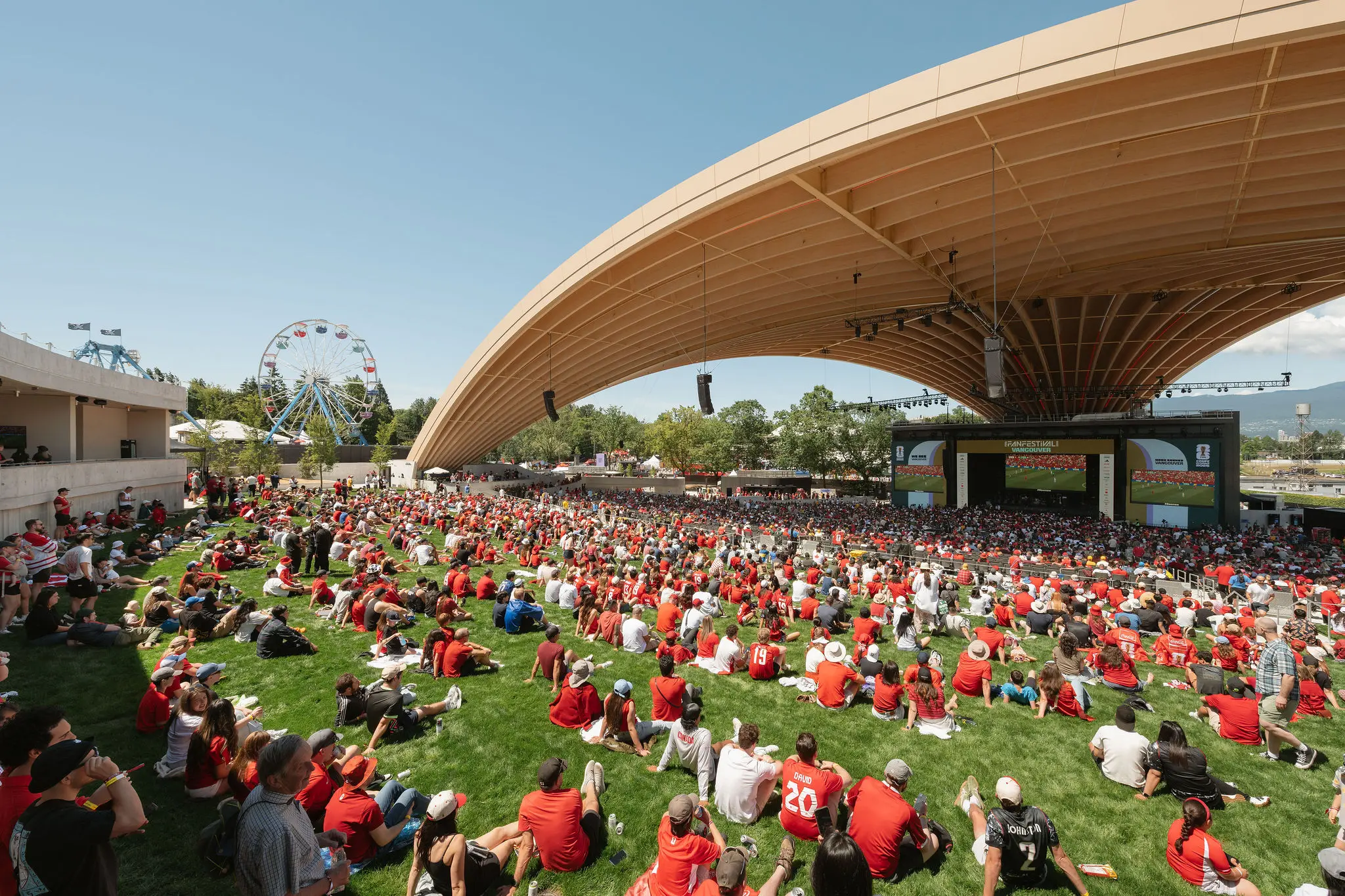 Large crowd wearing red sitting on grass under a curved wooden roof watching a large screen at an outdoor event with a Ferris wheel in the background.