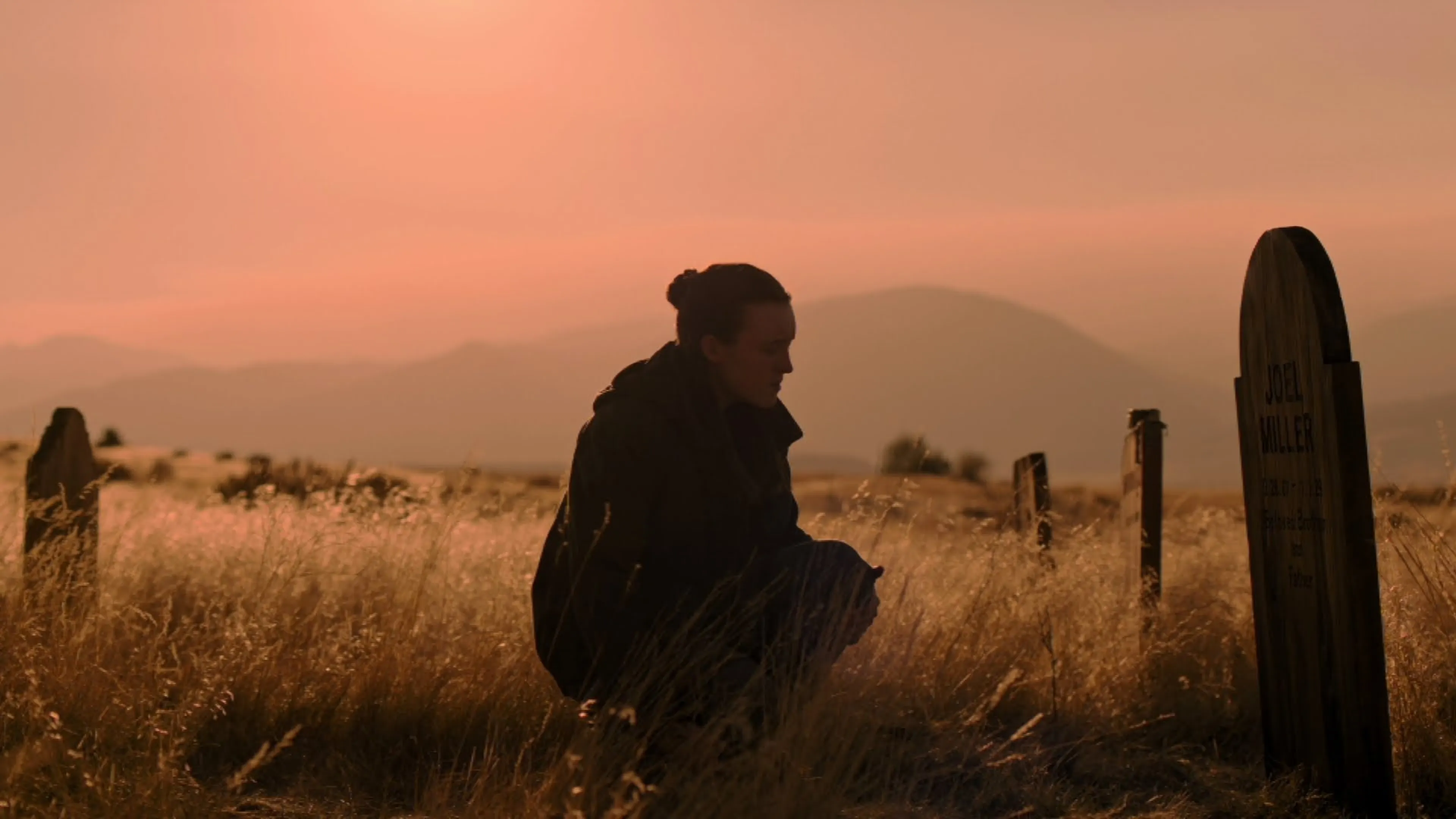 A person kneeling in a field of tall grass at sunset, facing a wooden gravestone with mountains in the background.
