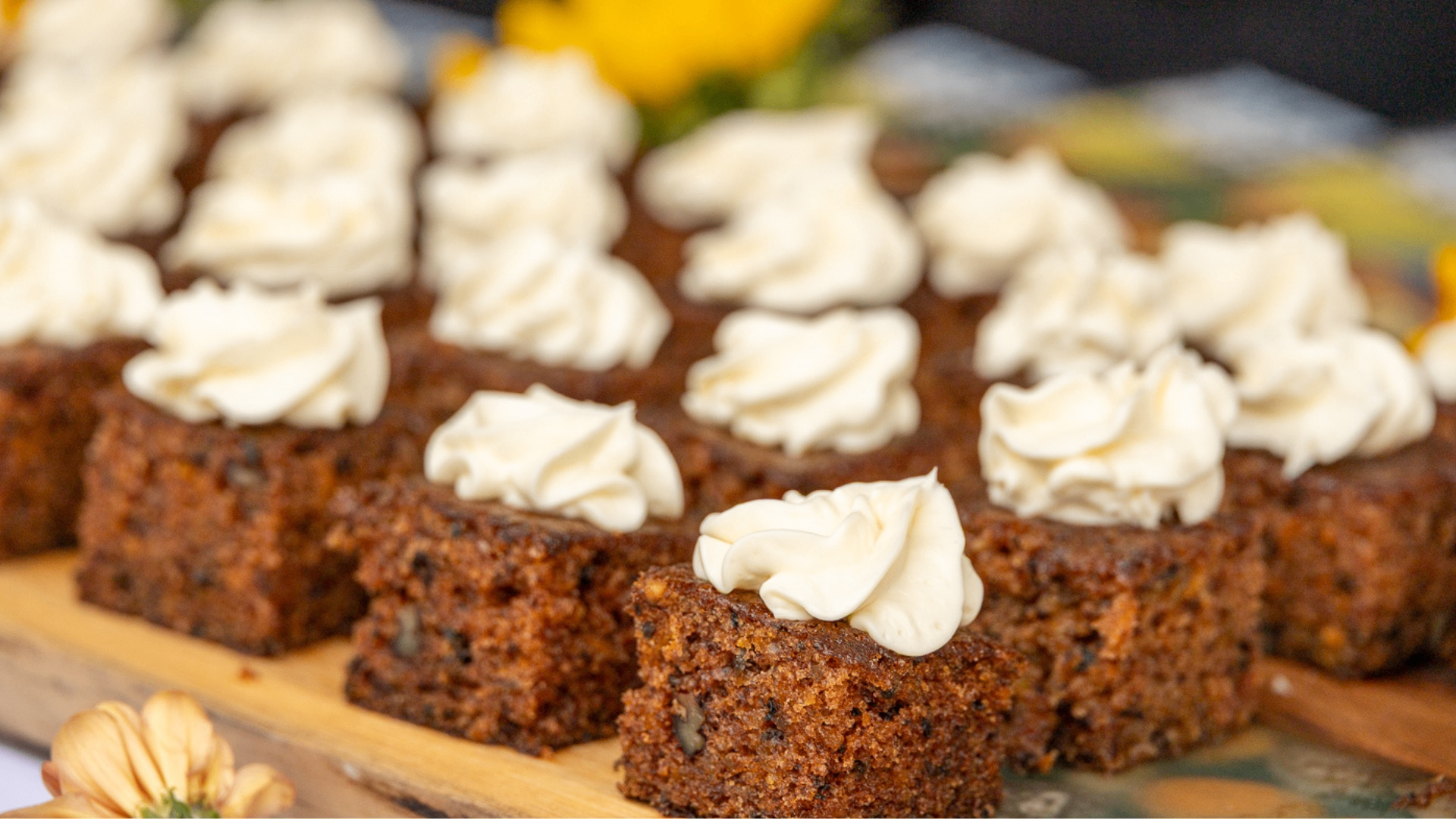 Rows of square carrot cake pieces topped with swirls of white frosting on a wooden board.