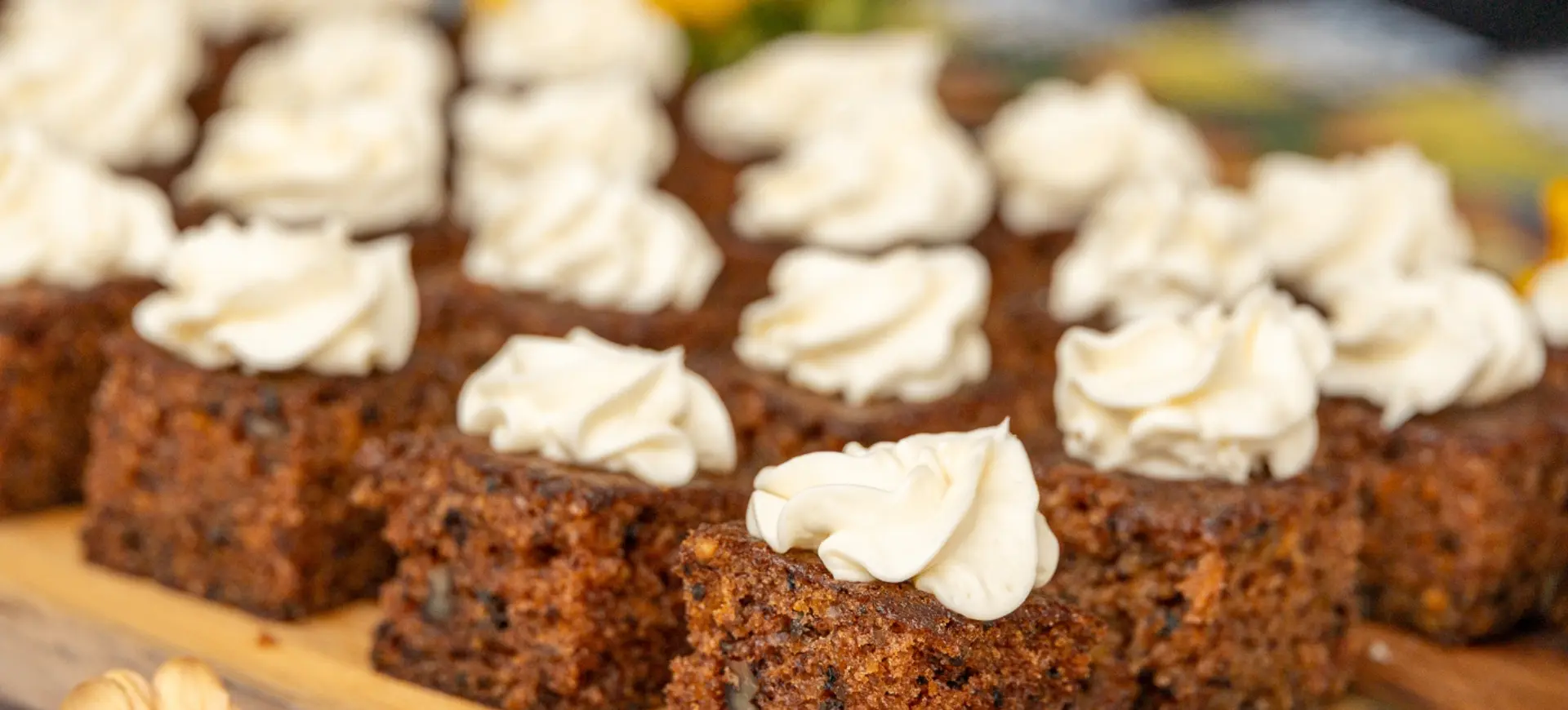 Rows of square carrot cake pieces topped with swirls of white frosting on a wooden board.
