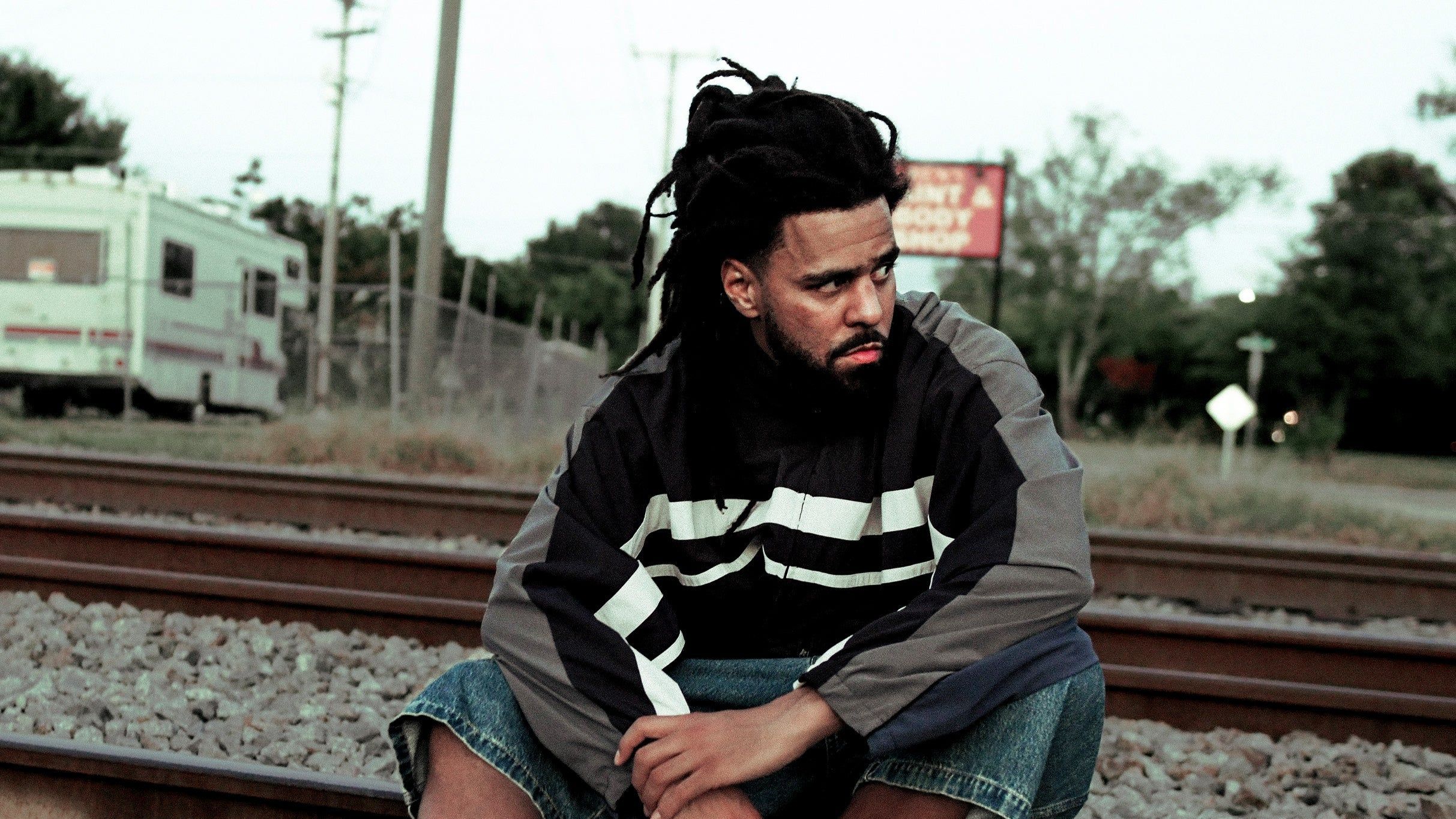 Person with dreadlocks wearing a black and gray jacket sitting on railroad tracks looking to the side.