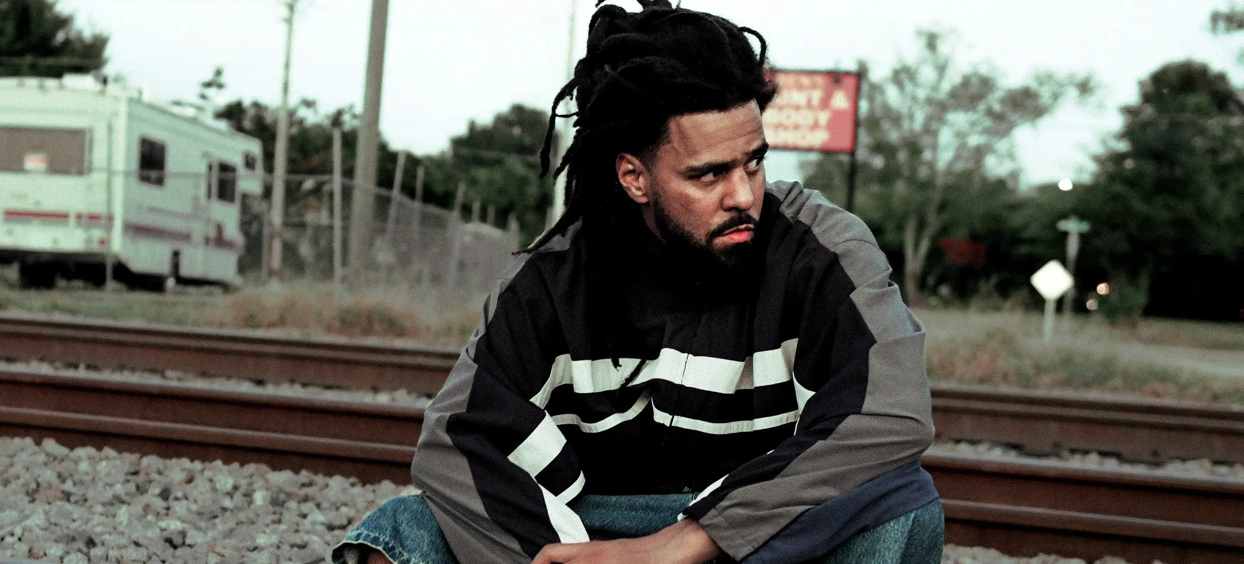 Person with dreadlocks wearing a black and gray jacket sitting on railroad tracks looking to the side.