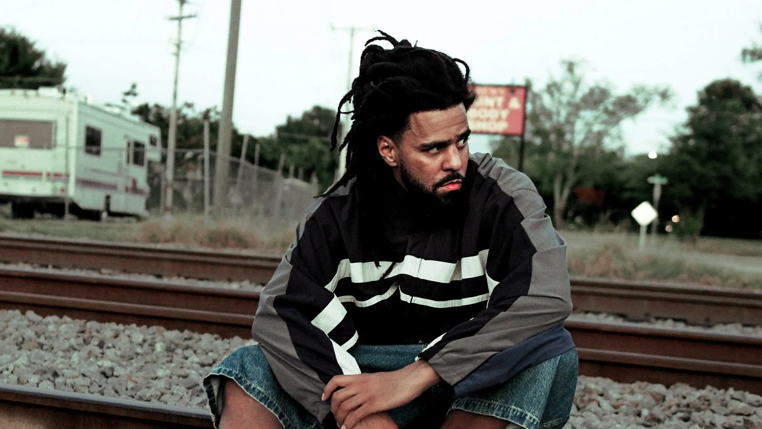 Person with dreadlocks wearing a black and gray jacket sitting on railroad tracks looking to the side.