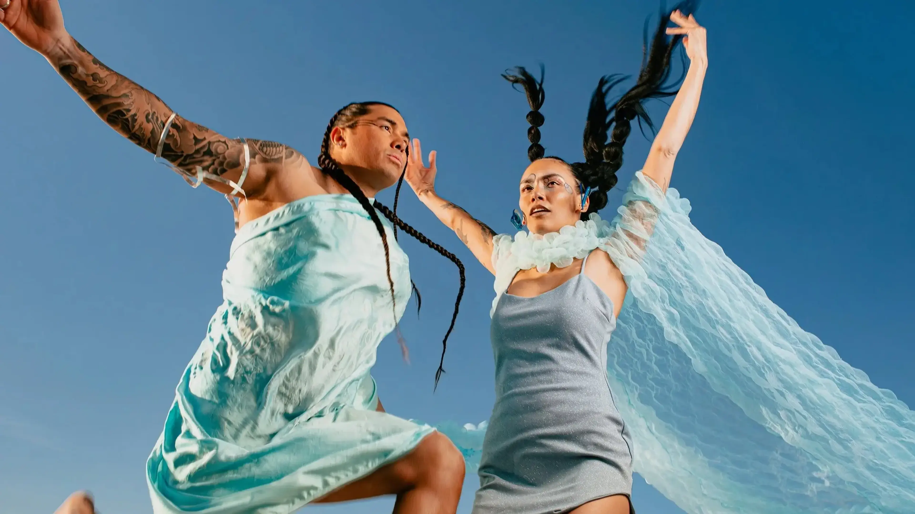 Two people in light blue dresses jumping with braided hair flying against a clear blue sky.