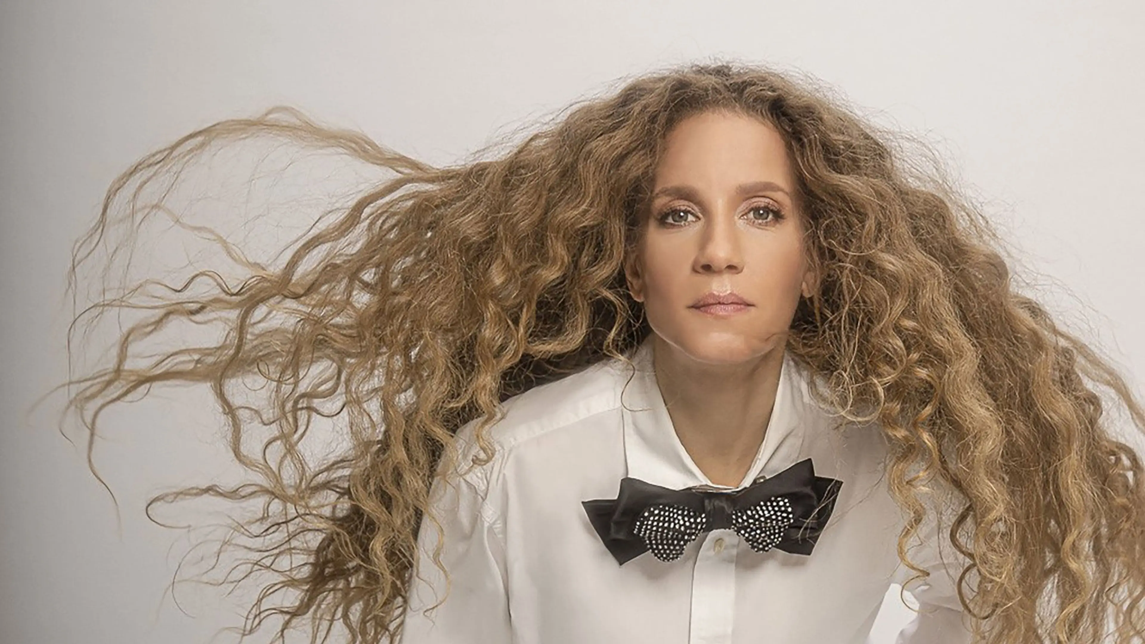 Person with long curly hair flowing sideways, wearing a white shirt and a black bow tie with rhinestones, looking forward.