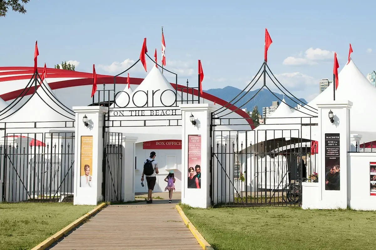Entrance gate with red flags and white tents labeled 'Board on the Beach' with two people walking inside.