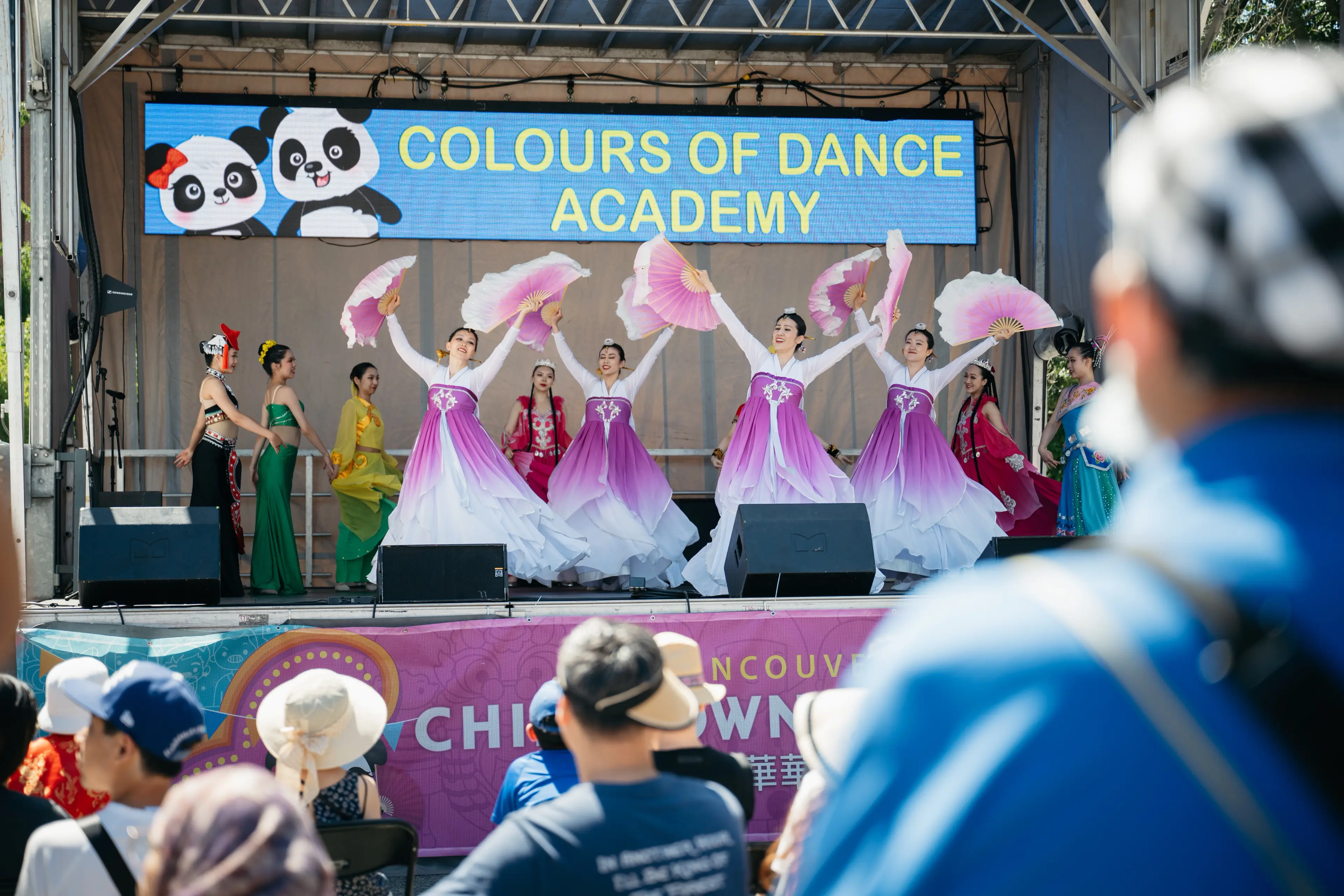 Group of dancers in colorful traditional costumes performing on stage with a sign reading 'Colours of Dance Academy' and an audience watching.