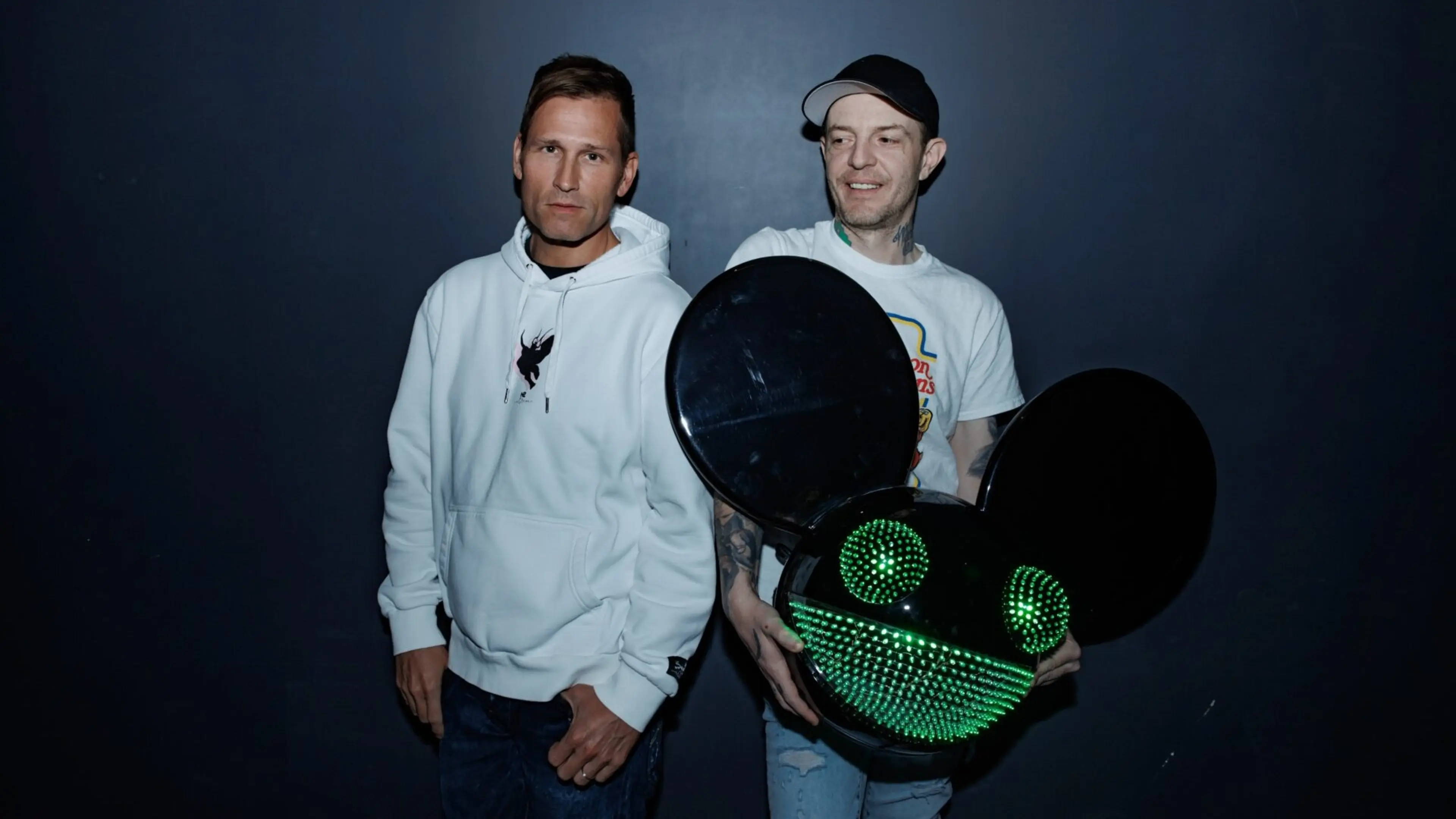 Two people standing against a dark background, one wearing a white hoodie, the other holding a large black helmet with green LED lights shaped like eyes and a smile.