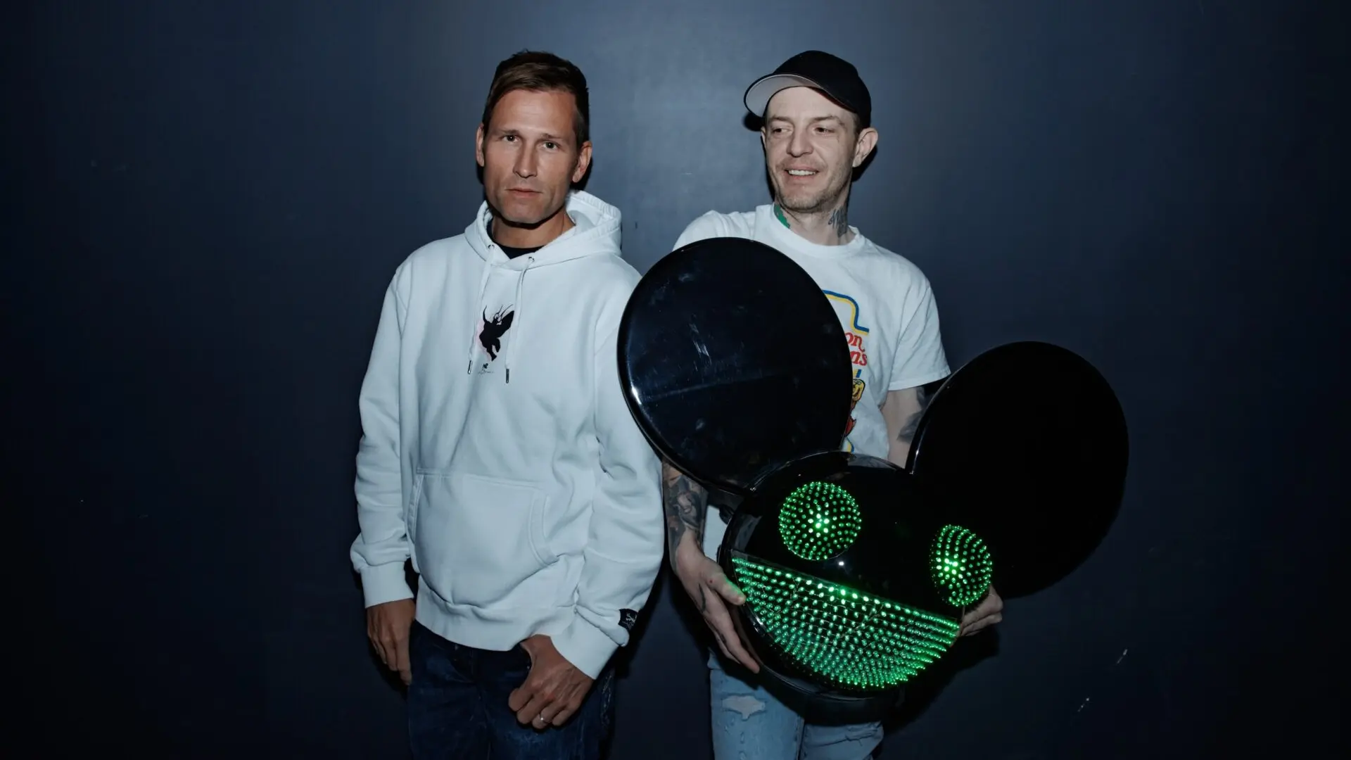 Two people standing against a dark background, one wearing a white hoodie, the other holding a large black helmet with green LED lights shaped like eyes and a smile.