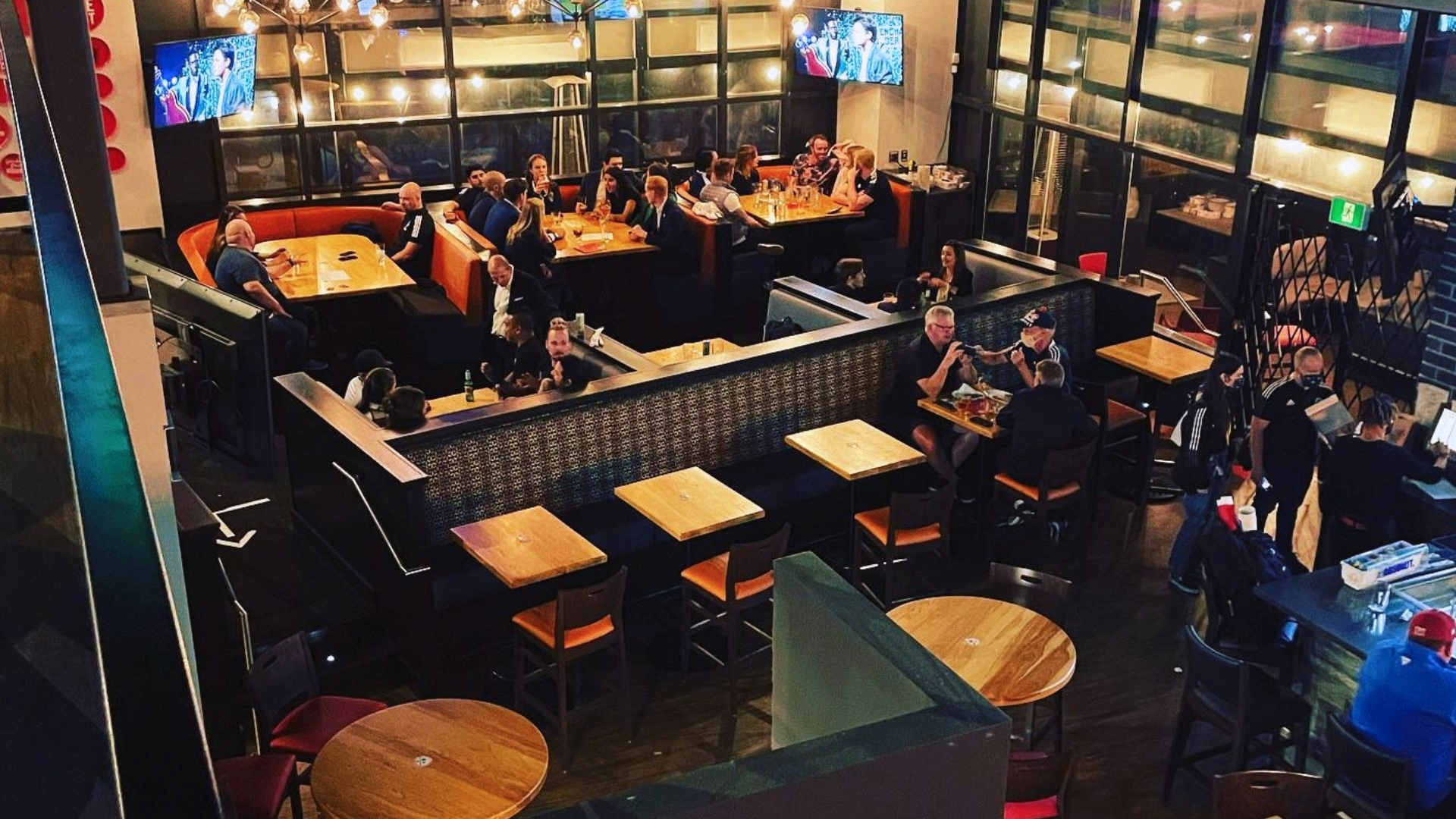 Interior of a busy restaurant with people sitting at tables and booths, two TVs mounted on the walls.
